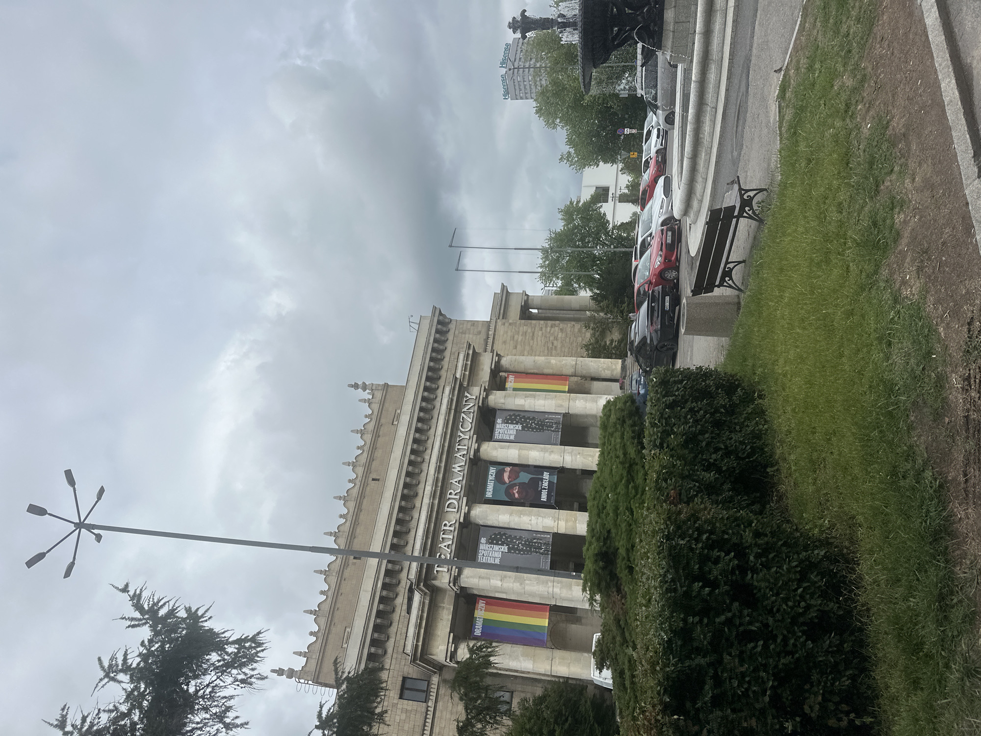 The image shows the Teatr Dramatyczny in Warsaw, Poland, on a cloudy day. The theater has a classical facade with columns and banners displayed. One banner features a rainbow flag, indicating support for the LGBTQ+ community. Other banners advertise upcoming shows. In front of the theater, several cars are parked in a lot. A green hedge and a lawn are visible in the foreground, with a park bench situated near the lawn. A fountain can be seen in the background. The overall scene is quiet and overcast.