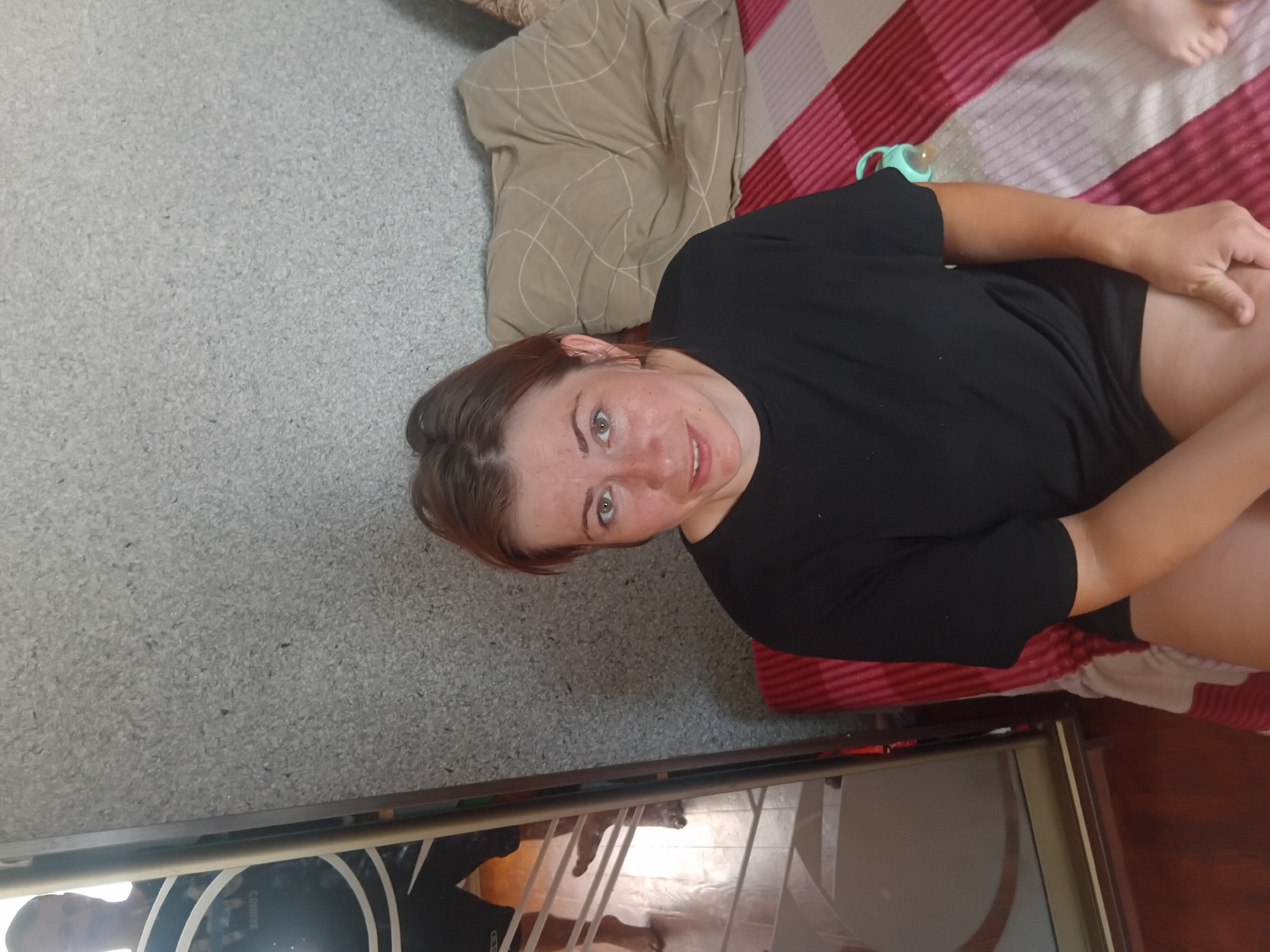 A medium shot shows a woman with auburn hair, freckles, and green eyes looking towards the camera. She is wearing a black t-shirt and black shorts. She is seated on a bed with a striped red and white comforter, a beige pillow, and a light blue pacifier with a white ring. To the left of the woman is a tall mirror that reflects a man wearing a black t-shirt and black shorts. The man's legs and feet are visible in the reflection. The background consists of a textured gray wall and a wooden floor. The scene is indoors, likely a bedroom, and the lighting suggests it is daytime. The location is not specified in the image itself.