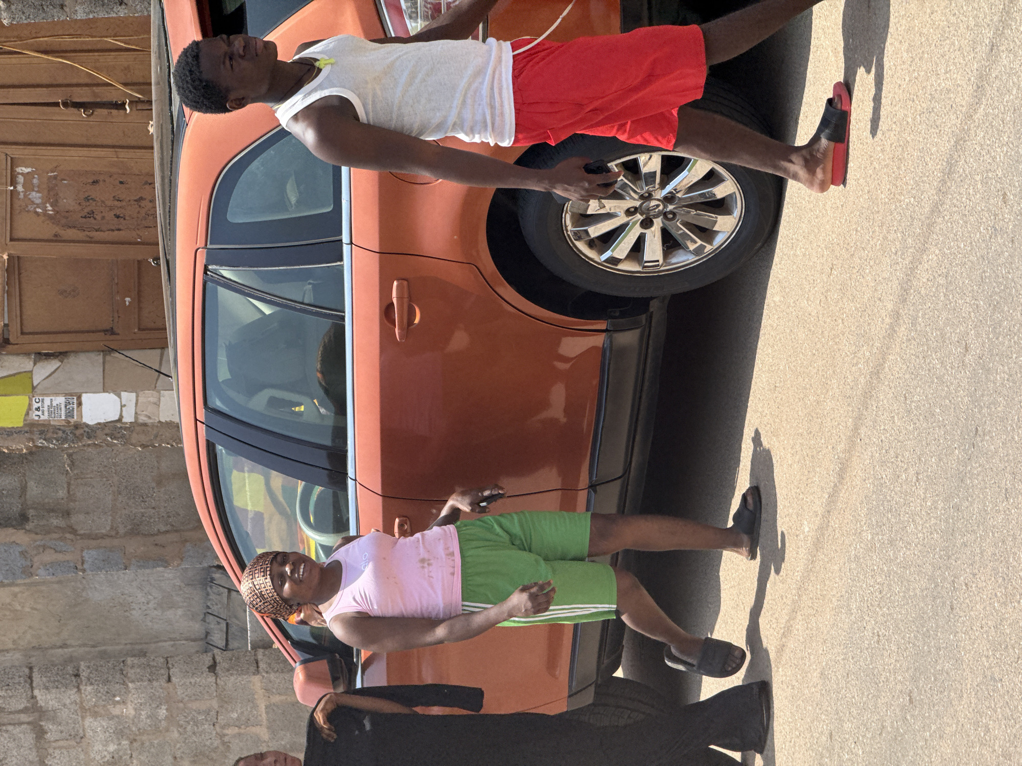 In Abuja, Nigeria, on a sunny day, three individuals are walking alongside an orange SUV parked on a paved street. The person in the foreground on the right is a young Black male wearing a white tank top and red shorts. He is looking to his right and has a necklace with a yellow pendant. To his left, a young Black female is walking, wearing a pink tank top and green athletic shorts with white stripes. She is smiling and looking towards the camera. She has a headscarf on. To her left, another individual, mostly out of frame, is wearing a black garment and appears to be walking away from the camera. The background shows a stone building with some graffiti and a wooden door. The tires of the SUV are visible, as are its chrome rims. The ground is a light-colored asphalt with shadows cast by the people and the car, indicating bright sunlight.