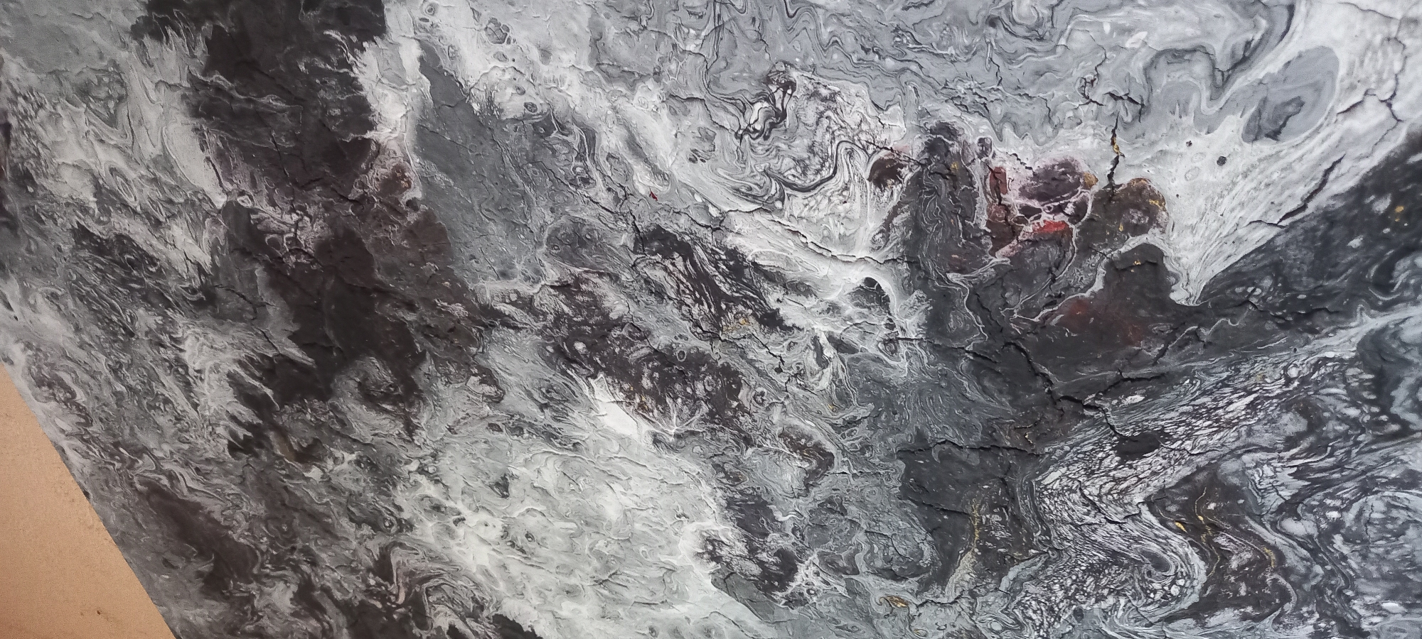 The image displays a close-up, overhead view of an abstract artwork or textured surface, confirmed to be located in Kryvyi Rih, Ukraine. The composition is dominated by a swirling, marbled pattern in various shades of black, dark grey, light grey, and white, with extensive crackling throughout. Fine networks of fissures and larger, irregular cracks fragment the painted surface, revealing darker underlying layers or recesses. Subtle specks of reddish-brown/maroon and metallic gold are visible within some darker areas, particularly towards the lower and right-center portions of the image. The upper-left corner features a smooth, unpainted, light brown/beige surface, indicating an edge or border of the artwork's substrate. The textured surface exhibits varied relief, suggesting a thick application of material or a complex drying process. No people, discernible objects other than the artwork itself, or specific actions are depicted. The environment is limited to the abstract surface and its immediate unpainted border.