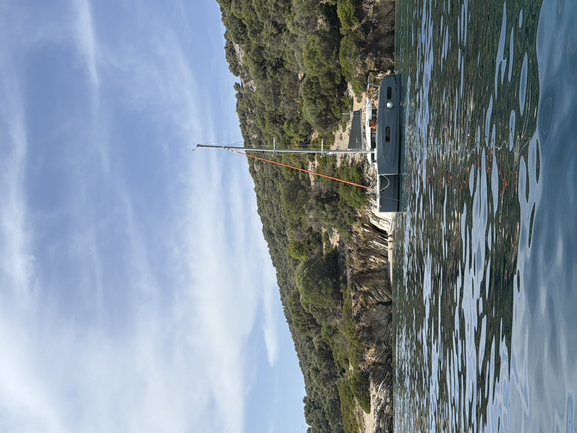 A gray catamaran is anchored in the calm, blue waters of a cove on the Municipal Unit of Kastos, Greece. The catamaran has two hulls and a tall mast with an orange rope running along it. The cove is surrounded by a steep, tree-covered hillside. Rocky outcrops and a small sandy beach are visible along the shoreline. The sky is a clear blue with a few wispy clouds. The water is slightly rippled, reflecting the sky and surrounding landscape. There are a few people on the beach in the distance.