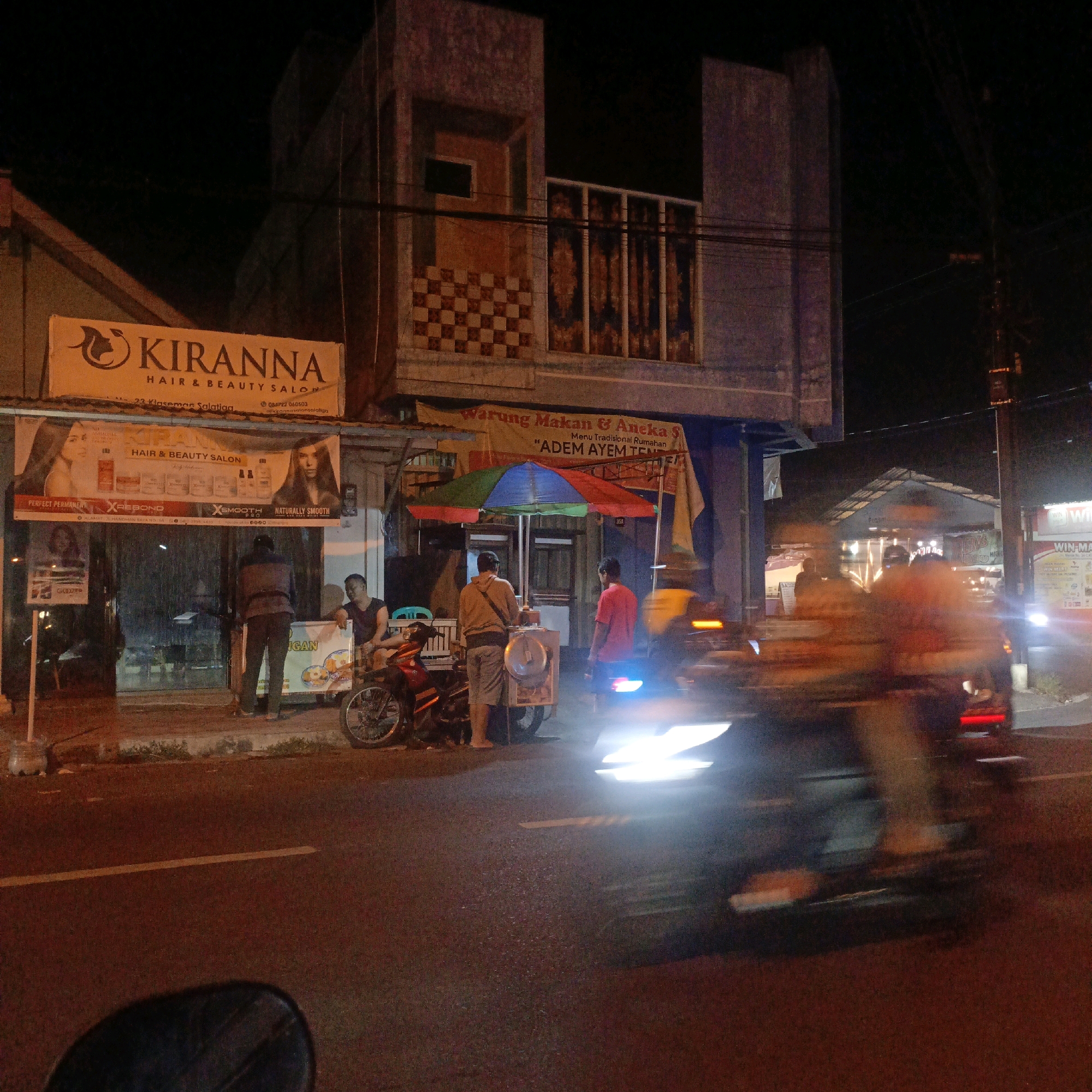 The image depicts a street scene at night in Salatiga, Indonesia. On the left side of the frame, a brightly lit sign for "KIRANNA HAIR & BEAUTY SALON" is visible, with additional banners below showcasing hair and beauty products. In front of the salon, a person stands near a motorbike. Further to the right, a food stall with a colorful umbrella is set up, attended by several people. One individual appears to be serving food from a cart, while others are standing nearby. The street is illuminated by streetlights and the lights from the establishments. Motion blur from passing vehicles is evident, suggesting active traffic. The overall environment is a bustling commercial area at night.