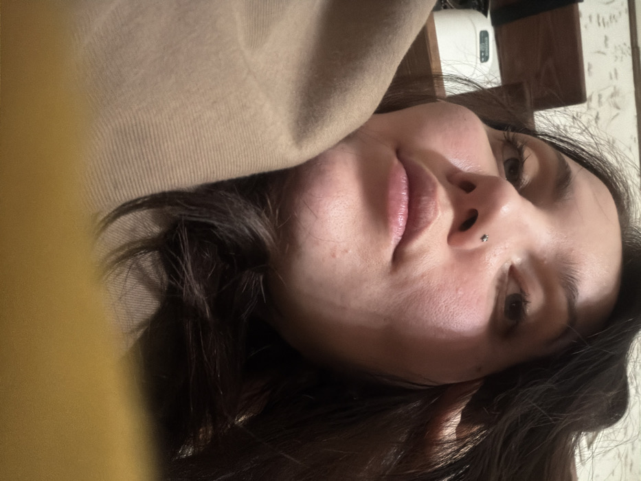 A young woman's face fills most of the frame. She has dark hair, visible mostly around her face, and fair skin. She has a nose piercing on one side. Her lips are full. She's looking slightly off-camera with a gentle expression. A soft, beige fabric drapes around her head. Parts of a room are visible in the background, including what seems to be wooden furniture and a white object.