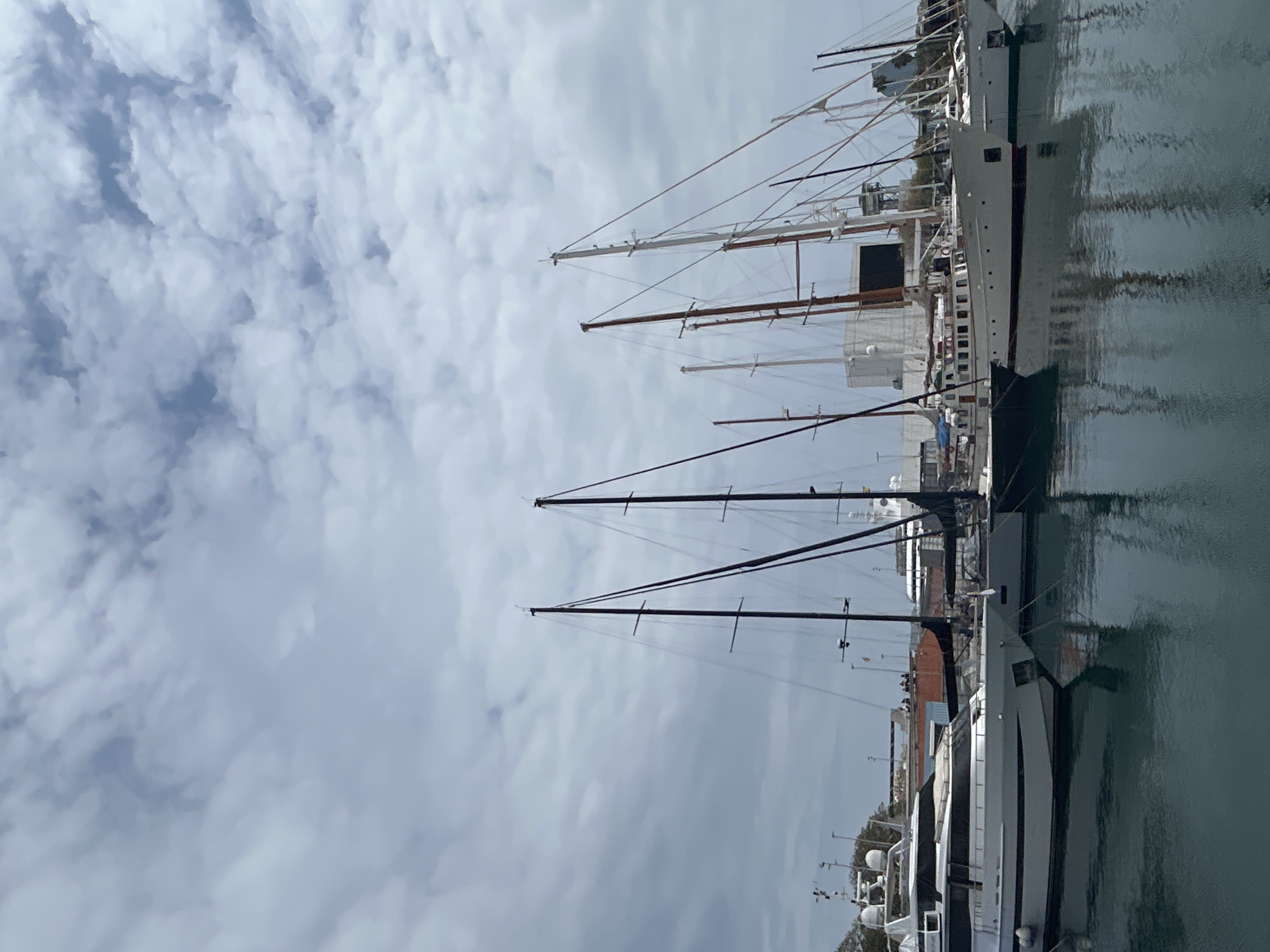 The image depicts a harbor scene in Barcelona, Spain. Several yachts and sailboats are docked, their masts reaching towards a cloudy sky. The vessels reflect in the water, creating a mirror image effect. The sky is overcast with textured clouds. Buildings and infrastructure can be seen in the background.