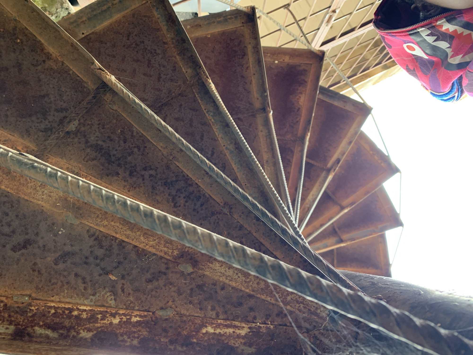This forensic image, captured from an extreme low-angle perspective, depicts the underside of a heavily rusted metal spiral staircase in Rafin Par, Nigeria, looking directly upward. The staircase structure is predominantly composed of dark reddish-brown, corroded metal plates forming the steps and a central support column, both exhibiting significant rust, pitting, and some flaking. Reinforcing bars (rebar), also oxidized, run parallel to the spiral, serving as structural elements or remnants of a railing system. Multiple cobwebs are visible across the rusted surfaces, indicating age and lack of maintenance. Bright white light from above illuminates the scene, casting the upper sections of the staircase in a darker silhouette. In the bottom right corner of the frame, the upper-right portion of an individual's head, obscured by a vibrant pink, purple, black, and white hooded sweatshirt with a distinctive graphic pattern (resembling a shark motif), is partially visible, indicating the presence of a person looking up at the structure. The environment beyond the immediate staircase structure appears to include vague geometric patterns, possibly from another building component or grating, situated against a bright, undefined background.