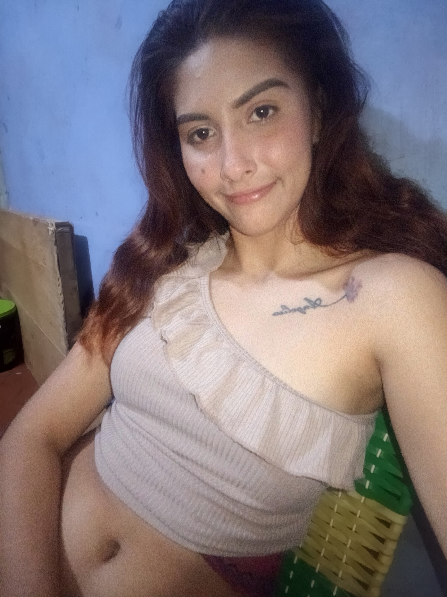 A woman is seated indoors. She has long brown hair and fair skin, and is wearing a light-colored one-shoulder top. The top reveals part of her abdomen. She has a tattoo on her upper left chest near her shoulder. She is sitting on a green and yellow chair. The background consists of a blue wall and part of what appears to be a wooden structure or cabinet. There is a green container on the left side of the image. There is no information available to determine the exact location in Juan Griego, Venezuela.