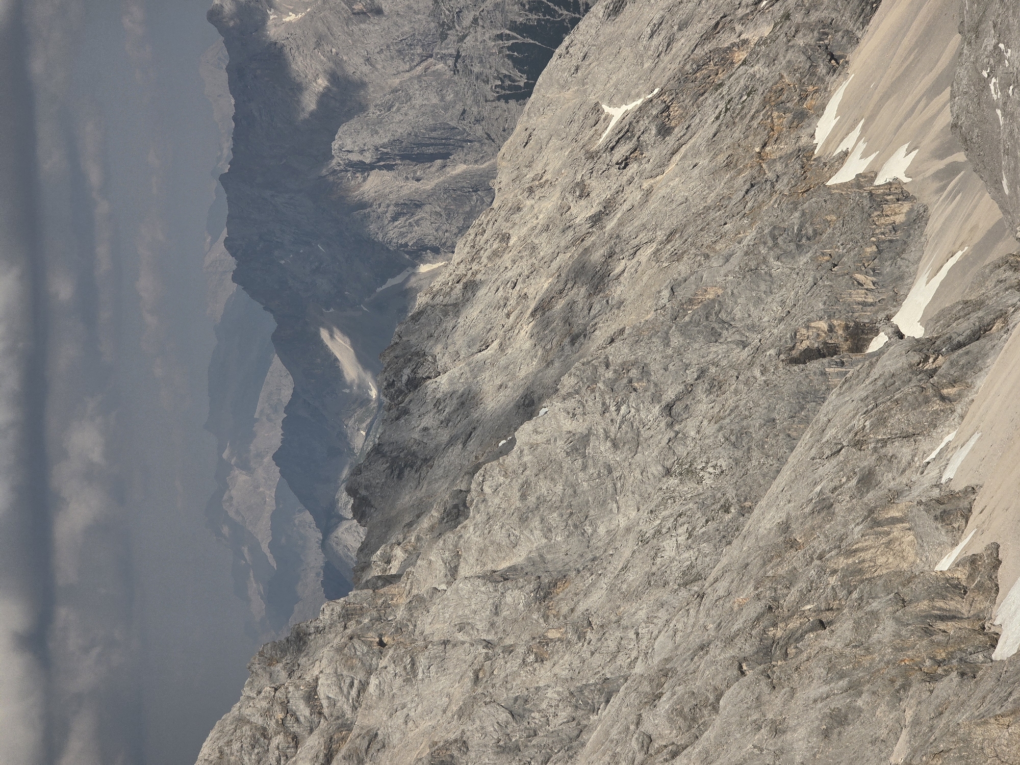 This media file depicts a dramatic, mountainous landscape, likely in the Garmisch-Partenkirchen region of Germany. The image is oriented vertically, presenting a steep, rocky incline that dominates the foreground and midground. The texture of the rock is rough and varied, with numerous fissures, ledges, and outcroppings. Patches of snow are visible in crevices and on some of the steeper slopes, suggesting a high-altitude environment or a recent snowfall.
In the background, layers of mountains recede into a hazy, atmospheric perspective. The distant peaks are shrouded in soft, grey clouds, which also drift across the left side of the image, obscuring parts of the mountain range. This cloud cover suggests possibly overcast or misty weather conditions, or it could be the early morning or late afternoon light casting shadows.
The scene is natural and wild, with no signs of human habitation or activity, such as buildings or infrastructure. The rugged terrain and the presence of snow indicate a remote, alpine setting, possibly popular for hiking or mountaineering, though no people are visible in the frame. The overall mood is one of grandeur and rugged natural beauty, with a sense of scale conveyed by the vastness of the mountains. The lighting appears to be diffused, consistent with cloudy conditions, and there is no visible text within the image.