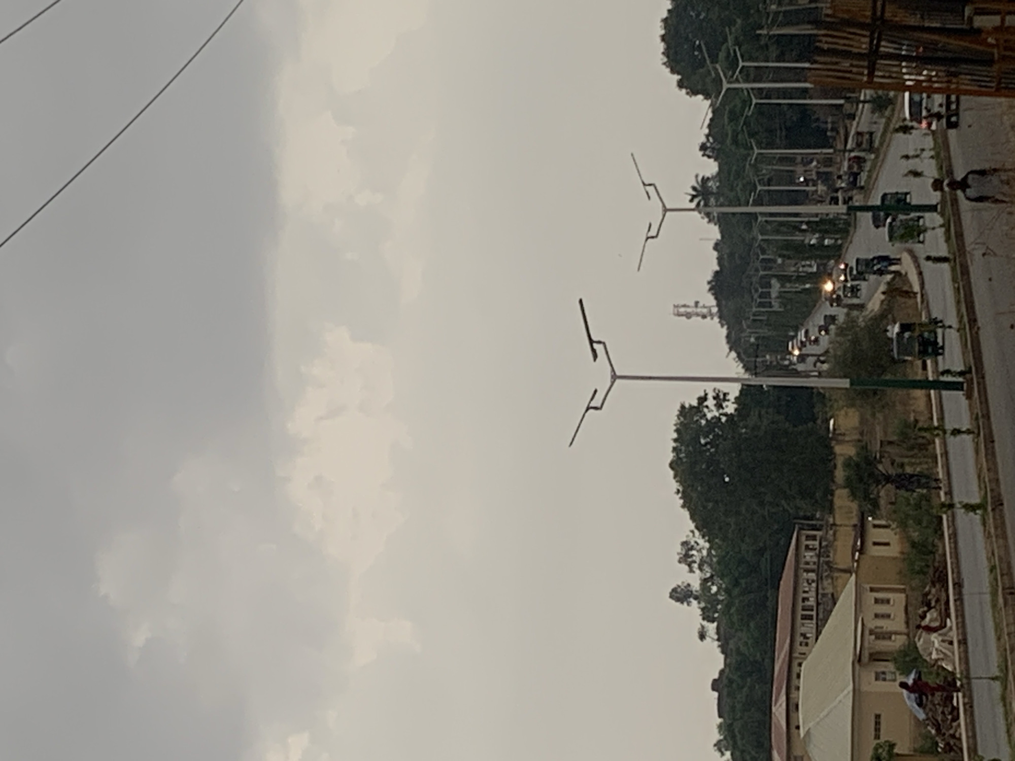 The image depicts a street scene in Jos, Nigeria under an overcast sky. The street is lined with modern streetlights and trees. A line of vehicles, including cars and tricycles, is visible along the road. A building with a red roof and multiple windows is situated on the right side of the street. Several individuals are present near the building and along the roadside. Construction materials or equipment are visible on the side of the road.