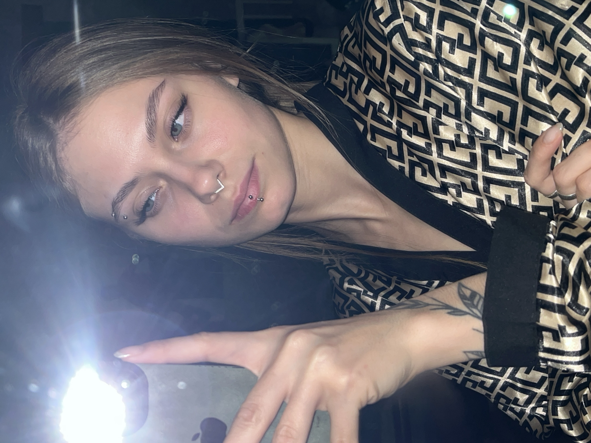 A young woman is holding a phone with the flash on. She is wearing a geometric patterned robe with black trim and has several facial piercings. A tattoo of leaves is visible on her forearm. The location of Фоки, Russia is not discernible from the image.