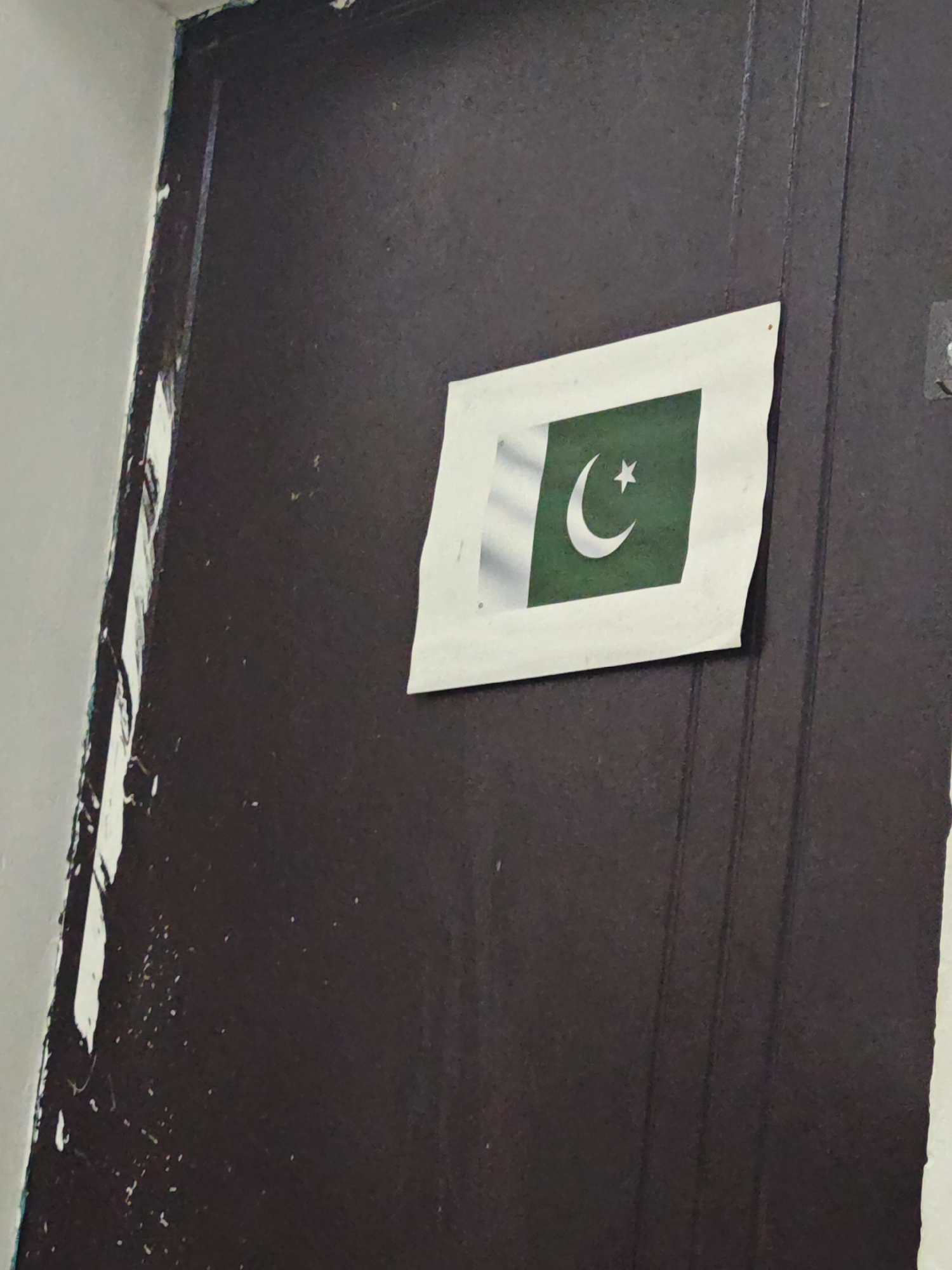 A Pakistani flag is taped to a dark-brown door. The door shows signs of wear and tear. It is not possible to infer its location or to make conclusions about the city of Chongqing High-tech Zone, China, from the visual information in the image. There are no people present in the image.
