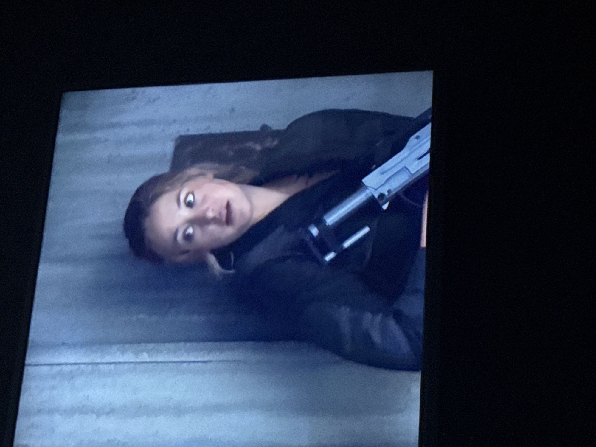 A woman with fair skin and light brown hair is lying on her side, angled diagonally in the frame. Her expression is one of surprise, with wide eyes and slightly parted lips. She wears a dark-colored jacket or coat. A gray and black weapon, resembling a futuristic gun, is held across her body. The background consists of a gray, textured surface, possibly a floor or wall. The location in the city of Mytishchi, Russia, cannot be determined from the visual information alone.