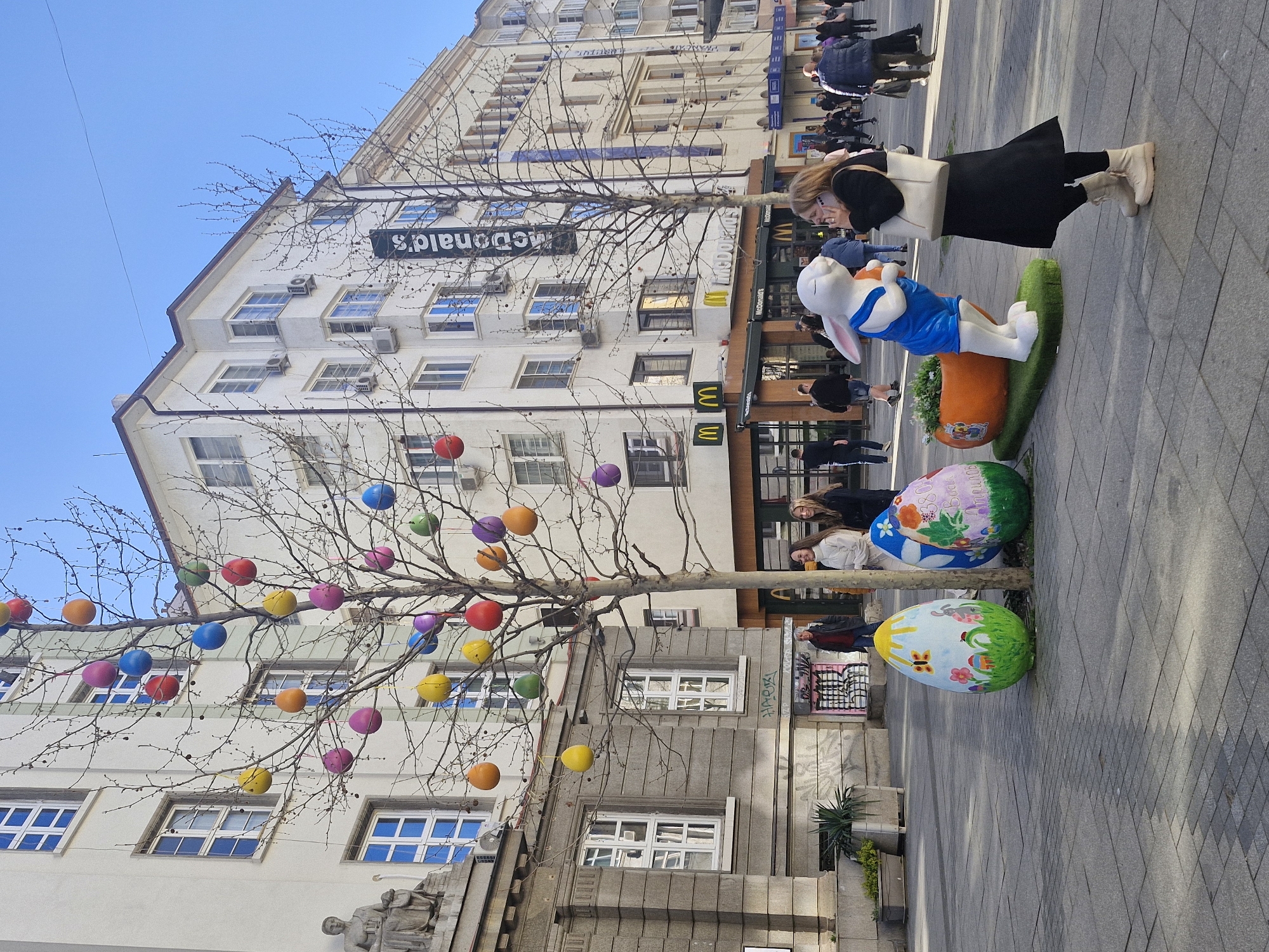 Captured at an outdoor location in Sofia, Bulgaria, the scene displays an urban setting with Easter decorations. Prominently featured is a bare tree adorned with colorful, round ornaments resembling Easter eggs. Adjacent to the tree are oversized, painted Easter eggs and a whimsical statue of a rabbit emerging from a carrot. A building with a McDonald's sign is visible in the background, with windows and air conditioning units. Pedestrians are seen walking in the background. A woman in a black dress and light-colored boots is positioned near the rabbit statue, appearing to interact with it, possibly taking a photo. The street is paved with grey tiles.