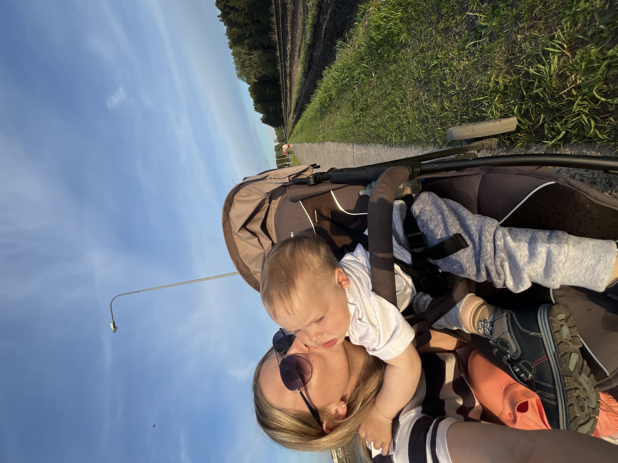 A woman wearing sunglasses and a striped shirt kisses a baby on the cheek. The baby is in a stroller, wearing a white shirt and grey pants. The stroller is on a paved path next to a grassy area and railroad tracks, under a blue sky with scattered clouds. A street lamp is visible in the background. The location is likely in the city of Chaikovsky, Russia.