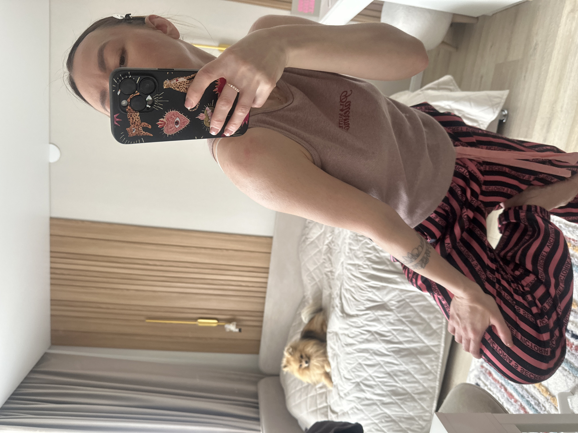 A woman is taking a selfie in what appears to be a bedroom. She is holding a smartphone with a patterned case. She wears a mauve tank top and black and pink striped pajama pants. A small, fluffy dog lies on the bed behind her. The room includes a bed with a white quilt, a wooden headboard, and a white nightstand. The location is unknown and the image does not contain information related to the city of Votkinsk, Russia.