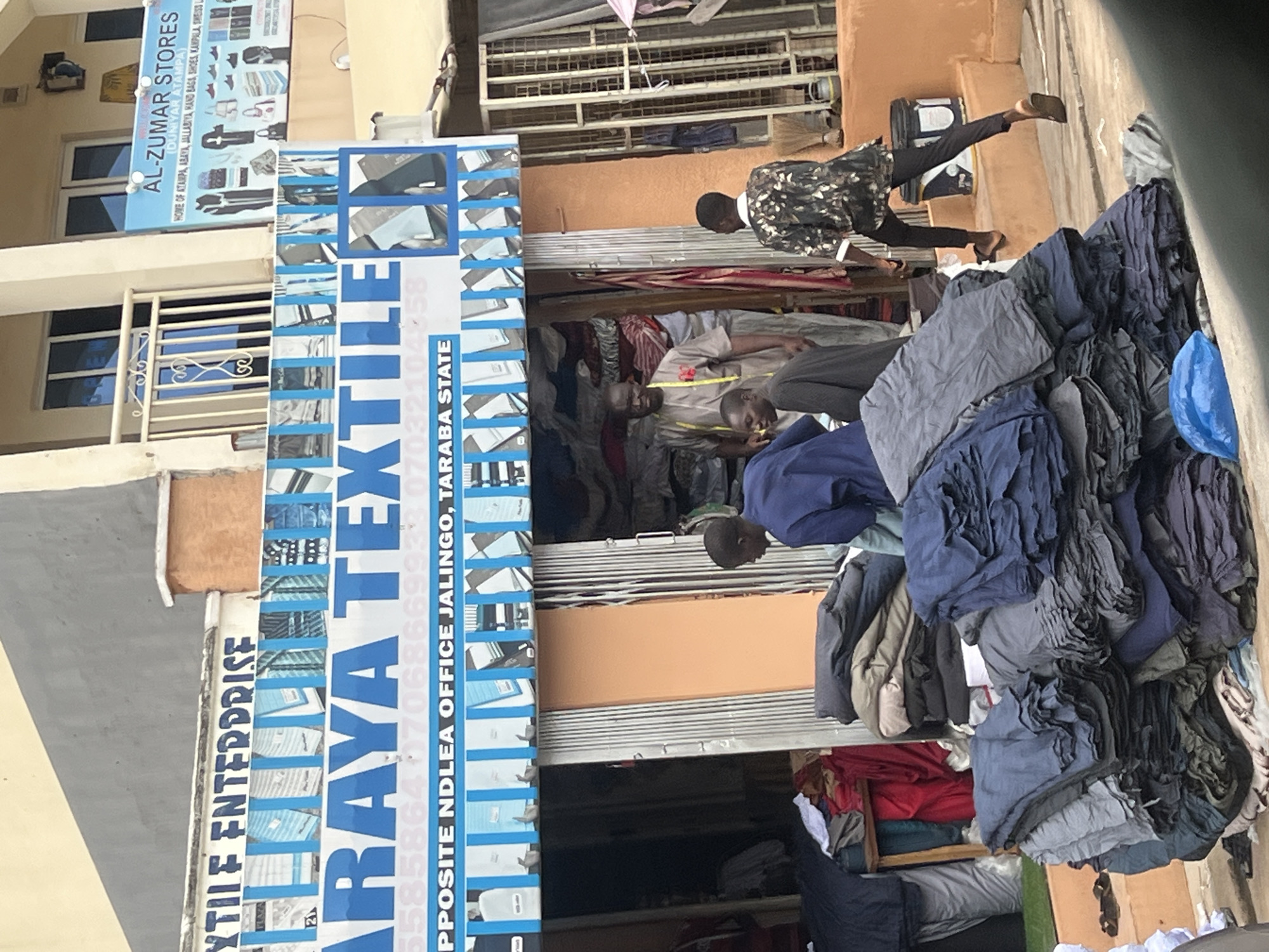 The image shows a street-level view of a textile shop named "ARAYA TEXTILE ENTERPRISE" located opposite the NDLEA Office in Jalingo, Taraba State, Nigeria. The storefront features a prominent blue and white sign with the shop's name and contact information. A collection of folded fabrics in various shades of blue and gray are piled high on the pavement in front of the shop. Several individuals are present, including a man in a tan garment with a yellow measuring tape around his neck, appearing to be a shopkeeper. Another man in a blue shirt is seen among the fabric piles, and a third person in a patterned shirt and black trousers is walking away from the shop. The shop's interior, visible through an open metal grate door, contains more textiles. To the right of the textile shop, a sign for "AL-ZUMAR STORES" is visible, advertising various clothing and accessories. The environment suggests an active commercial area in Jalingo.