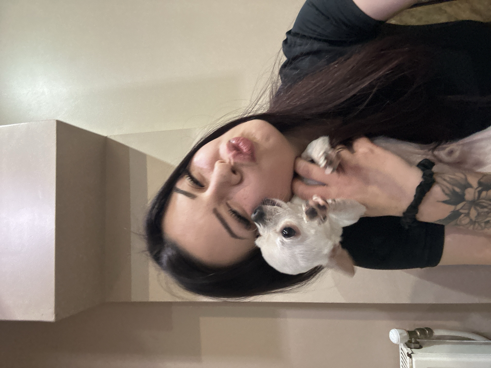 A woman with long dark hair is holding a small, white Chihuahua. She has her eyes closed and is puckering her lips as if to kiss the dog. The woman is wearing a black top and has a tattoo on her arm. A black hair tie is on her wrist. The background consists of a neutral-colored wall and trim, and a radiator is visible in the lower right corner. The location is unidentifiable as Sliven, Bulgaria.