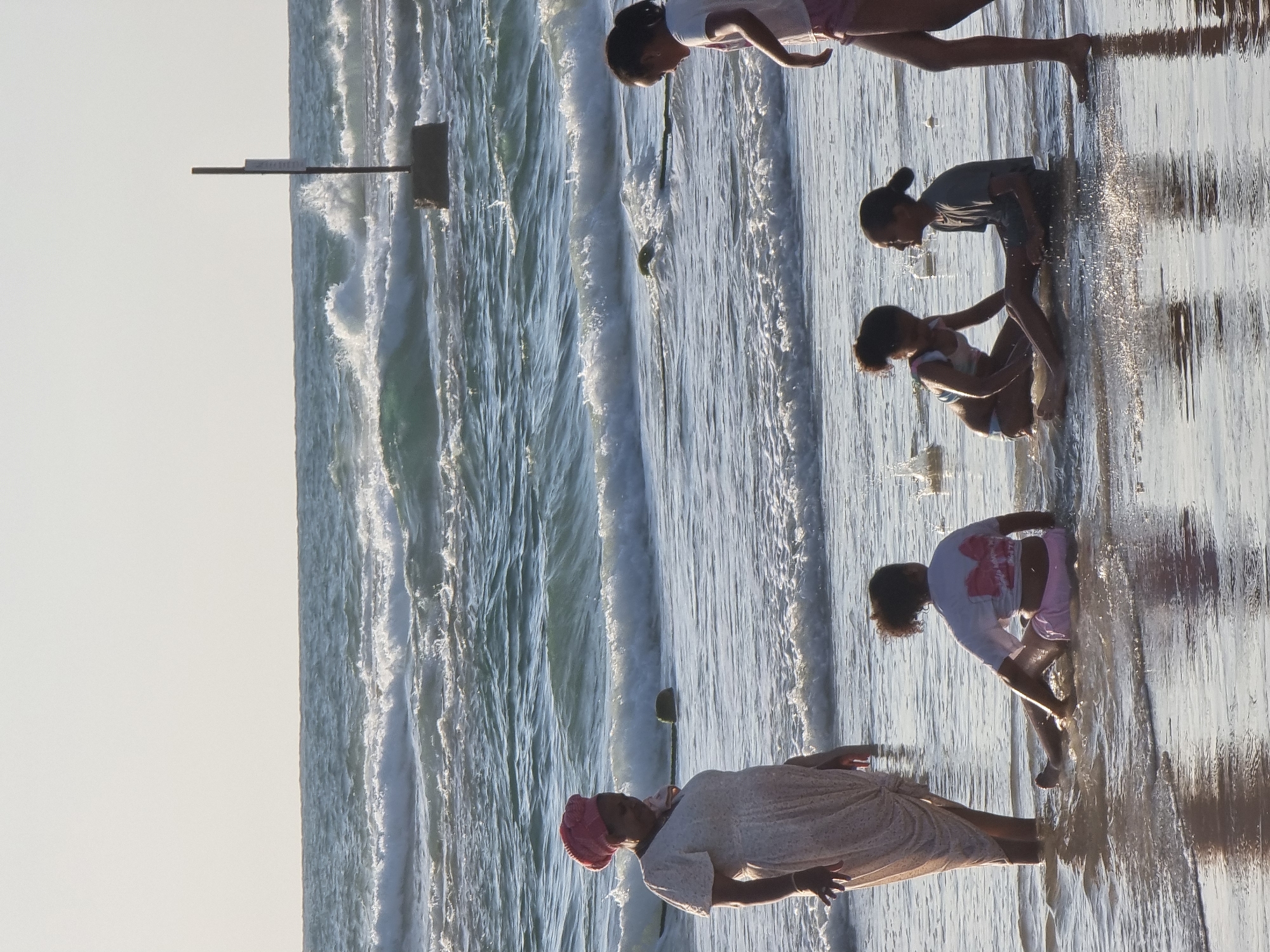 Captured at an eye-level perspective, the image portrays individuals enjoying the beachfront of Tel-Aviv, Israel. On the right side of the scene, five individuals appear to be playing in the water's edge. The first figure is walking through the water, the second and third are crouched down, and the fourth and fifth are standing in the water. The ocean waves flow from the left side of the frame towards the right, leading to the shoreline. A lifeguard station, consisting of a metal structure, is visible on the left, slightly farther away from the individuals playing in the water. The sky above is a pale shade, possibly suggesting it is either early morning or late afternoon.