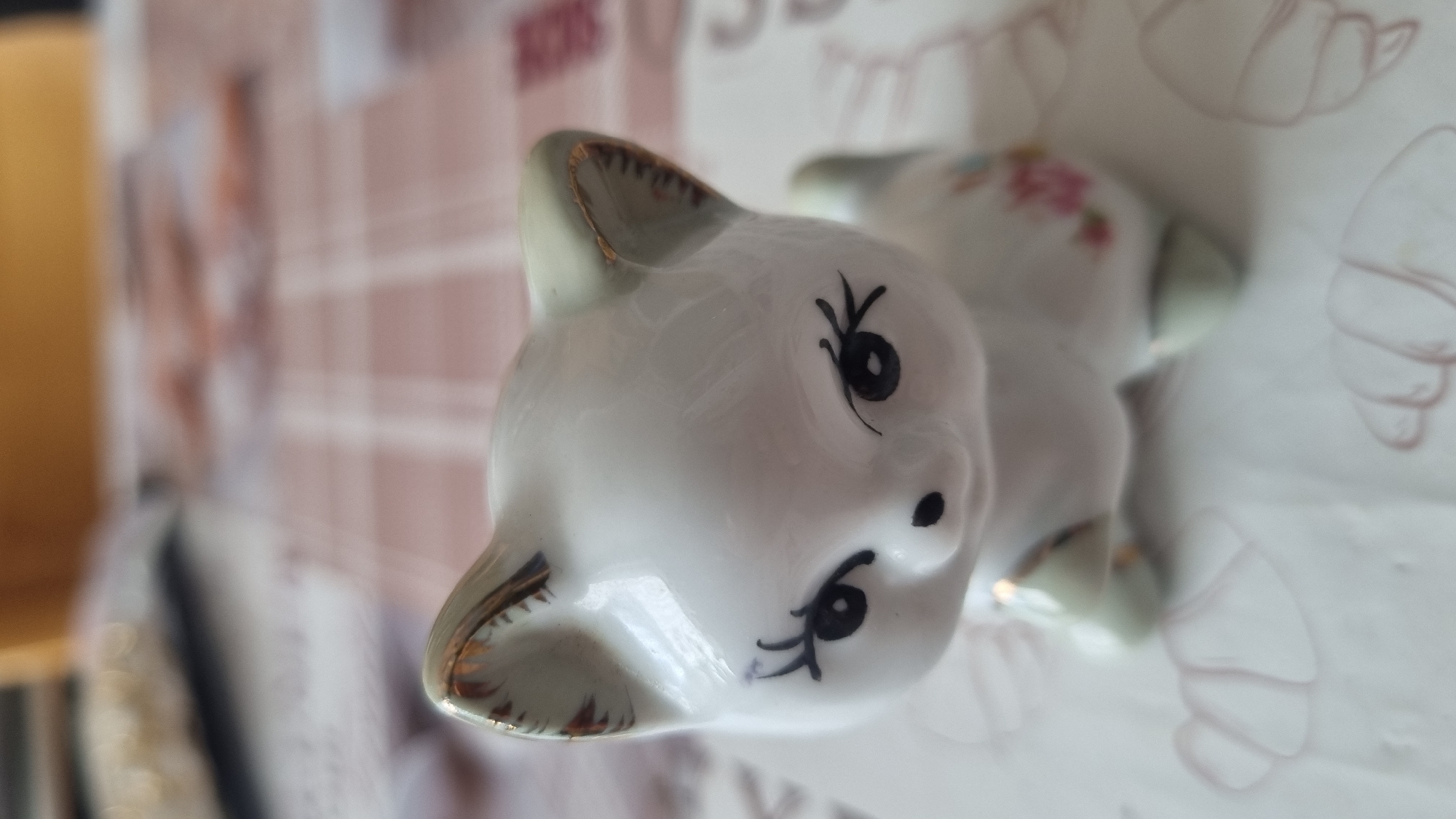This is a close-up, eye-level, indoor photograph of a small, white ceramic cat figurine. The figurine is depicted from the chest up, with its head tilted slightly forward. It has large, black, painted eyes with long, delicate eyelashes. Its nose is a small, black dot. The inside of its ears and the edges of its ears have a gilded trim, with some chipping visible on the right ear. The body of the cat has subtle floral patterns in pink and blue. The figurine is placed on a white surface with faint, light pink line drawings of croissants. The background is blurred, showing a checkered tablecloth in shades of pink and white, with a blurry yellow object at the top. The image does not provide any information or visual cues that would allow for its location to be identified as Smila, Ukraine.