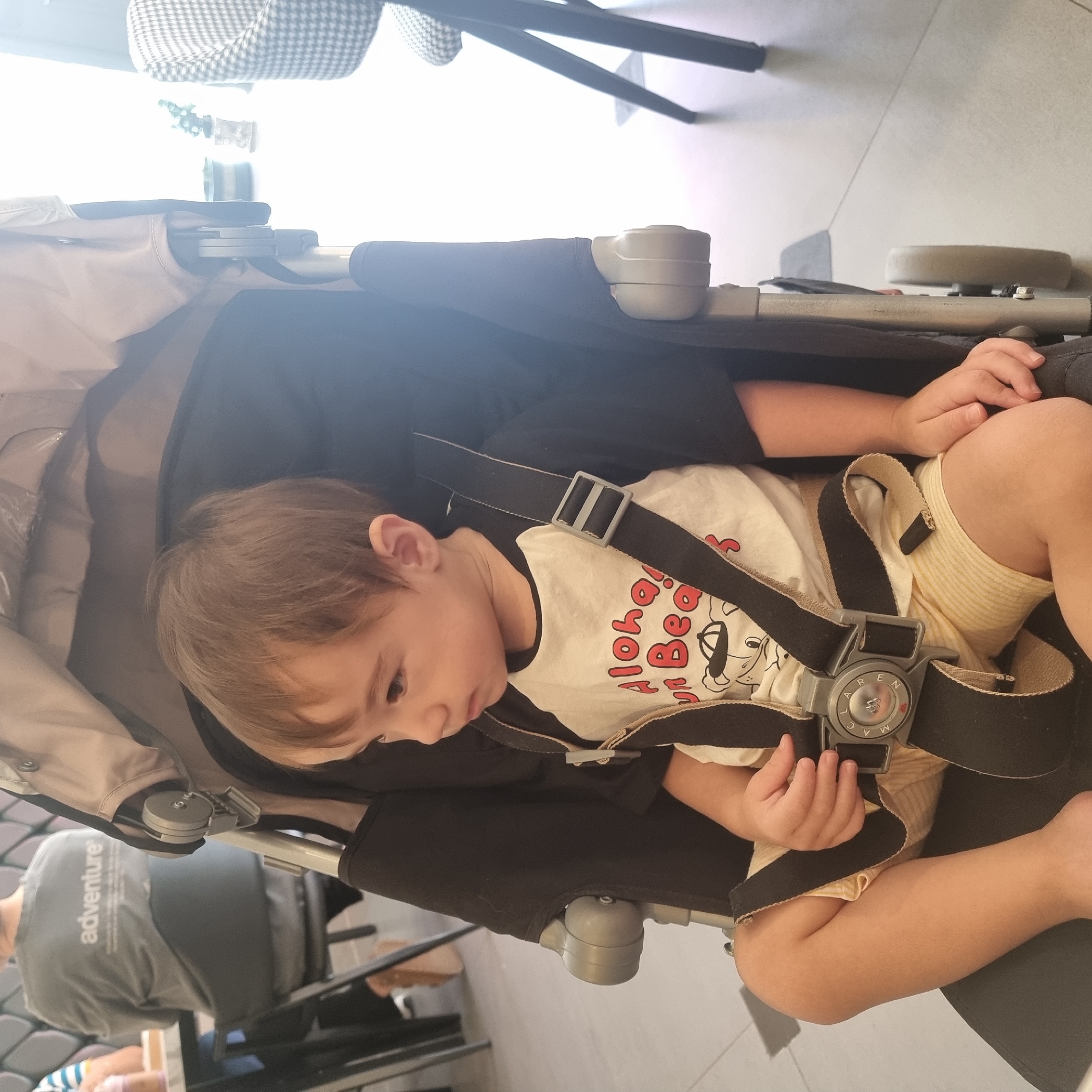 A young child with light brown hair is seated in a stroller, facing to the left of the frame. The child wears a t-shirt with black sleeves and a white front featuring red lettering that reads "Aloha" and "Bear," along with a cartoon character graphic. Yellow striped shorts are also visible. The stroller has a grey frame and black fabric, with a five-point harness securing the child. The background is blurred, showing a person in a grey t-shirt with "adventure" printed on the back, sitting at a table. To the right, a chair with a black and white patterned upholstery is visible. The floor appears to be light-colored tile.
