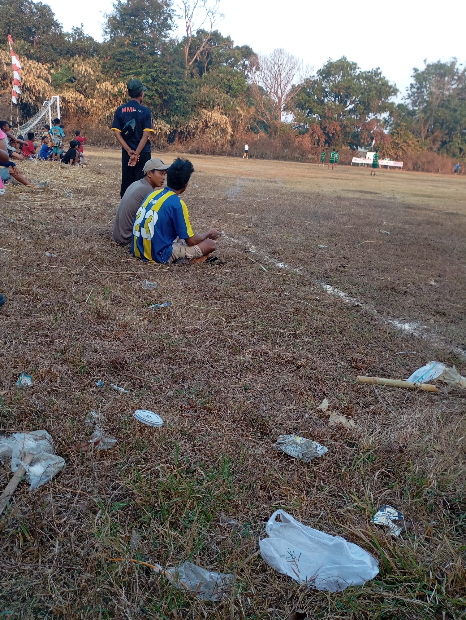 In the city of Kecamatan Haurgeulis, Indonesia, a football match is in progress on a dry, brown field marked with a white line. In the foreground, the field is littered with various types of trash, including plastic bags, wrappers, and a bamboo stick. Three young men are sitting on the ground, facing away from the camera and towards the game. The man in the middle wears a blue and yellow jersey with the number 23. Behind them, a man in a dark shirt with "MMR 2014" written on the back stands facing the field, his hands clasped behind him. Further in the background, several children are gathered near a soccer goal, and players in green jerseys are scattered across the field. The scene is framed by trees and foliage in the background, with a clear sky overhead.