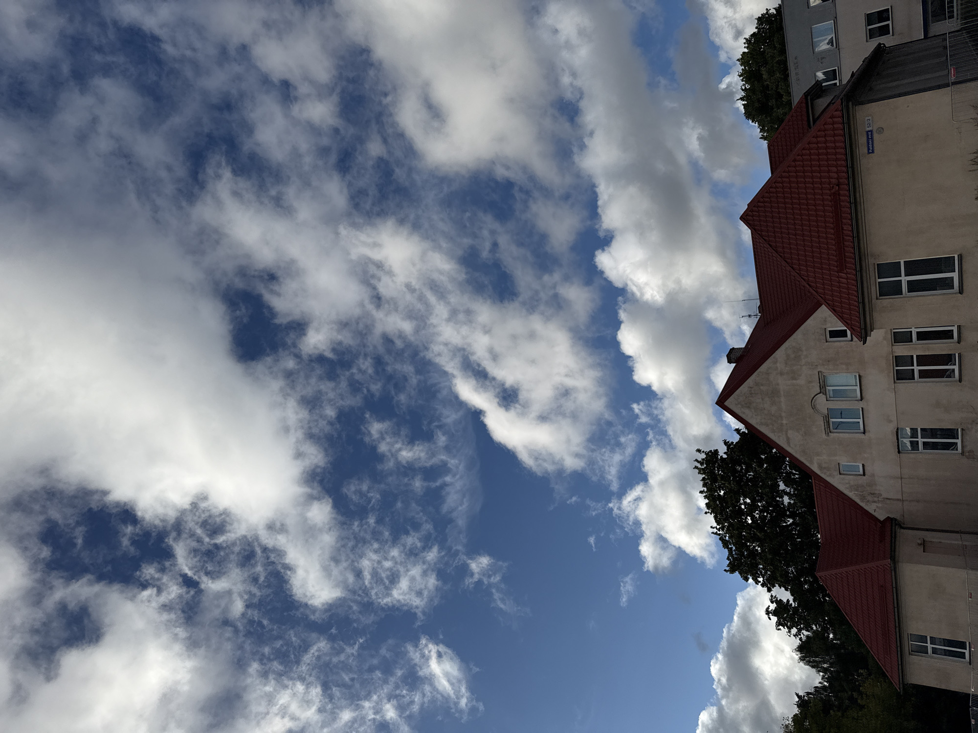 This is a photograph taken in Tallinn, Estonia, featuring a clear blue sky with scattered white clouds. The image is oriented vertically, with the buildings and trees appearing to the right side of the frame. The architecture suggests a residential or commercial area, with multiple windows visible on the buildings and red-tiled roofs. Trees are present, partially obscuring some of the buildings and contributing to a natural element within the urban setting. The lighting indicates it is daytime, likely with a bright sun given the clear sky. No people are visible in the image. A street sign with text "Pudisoo tee 52/2" is visible on one of the buildings.
