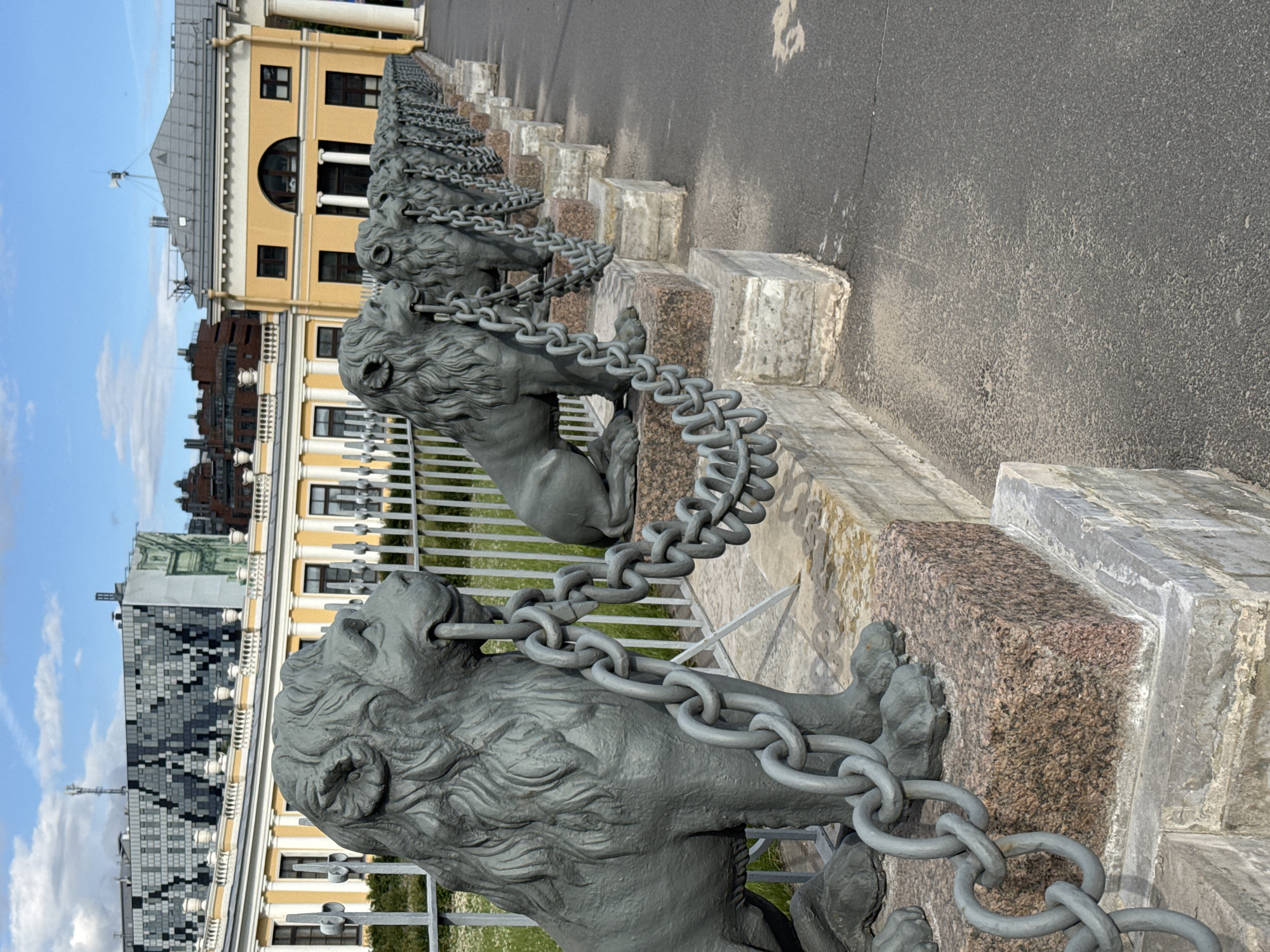 This media file captures a striking visual of a chain fence adorned with lion sculptures, set against a backdrop of classical architecture and a clear blue sky dotted with clouds. The scene is oriented vertically, with the ground and road appearing on the right side of the frame. The foreground is dominated by two intricately sculpted grey lion statues, each with a thick, heavy chain held in its mouth, linking them together. These sculptures are positioned atop stone pedestals, with the one in the foreground being a mix of grey and pink granite, and the one behind it appearing to be a lighter grey stone.
In the midground, a series of similar lion sculptures and chains recede into the distance, forming a continuous barrier. Behind the fence, a pale yellow classical building with a grey roof and arched windows is visible. Further back, a darker, modern building with a patterned facade of grey and black squares stands out. The overall setting suggests an outdoor public space, likely a park, promenade, or the grounds of a significant building in Saint Petersburg, Russia.
The lighting indicates it is daytime, with the sun casting subtle shadows. The weather appears pleasant with a bright sky. The emotion evoked by the scene is one of grandeur and strength, with the powerful lion sculptures symbolizing protection or dominion. There is no visible text in the image. The detailed sculpting of the lions and the heavy chains are notable details that contribute to the imposing nature of the fence.