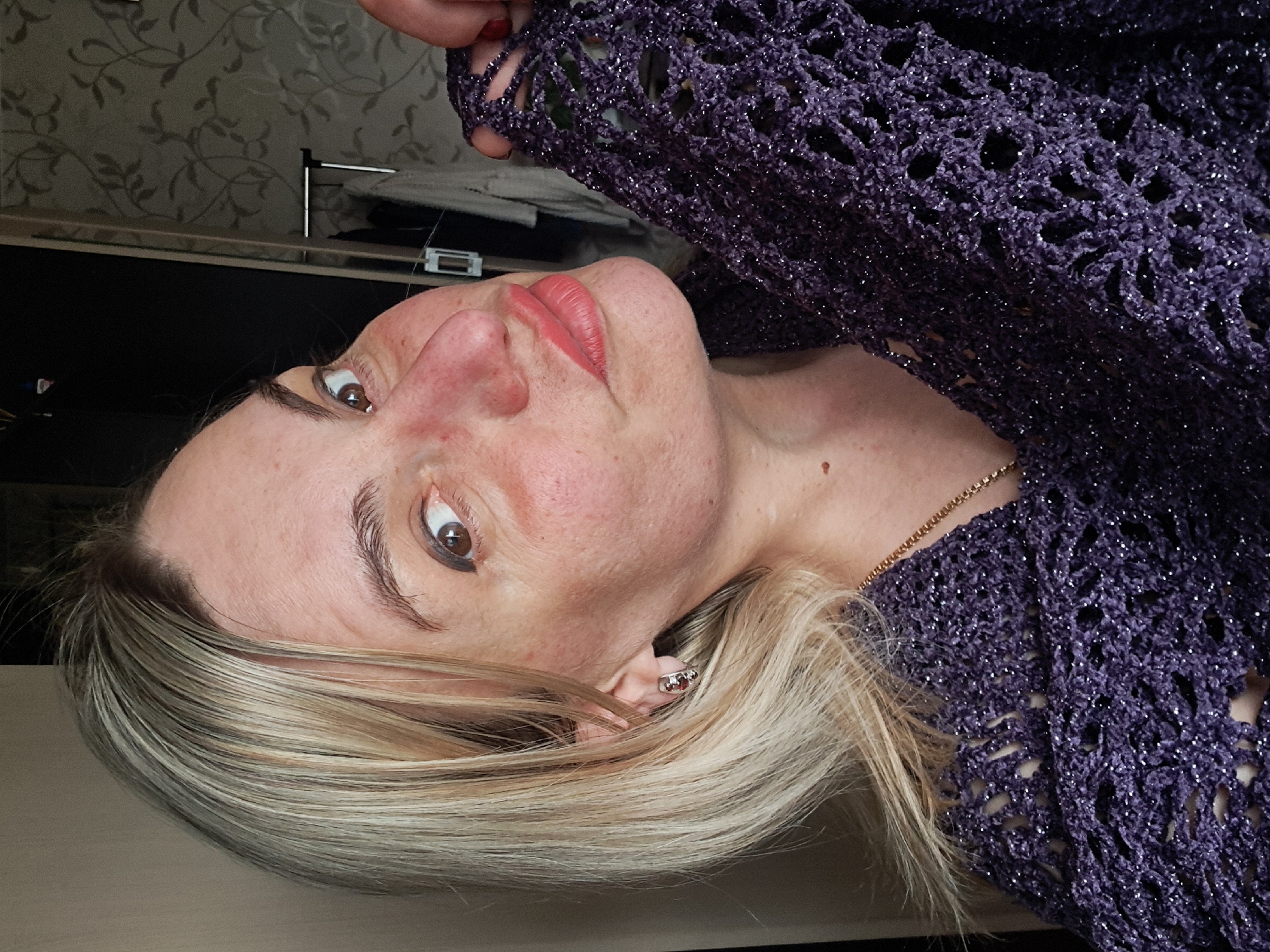 This close-up portrait shows a woman in an indoor setting. The main subject is a woman with blonde hair, styled around her face, and dark brown eyes looking directly at the camera. She has fair skin, with some redness visible on her cheeks and nose, and her lips are painted with pink lipstick. A small earring, possibly with red stones, is visible in her right ear, partially obscured by her hair.
She is wearing a dark purple, intricately crocheted garment, possibly a shawl or sweater, which features glittering threads that catch the light. A delicate gold chain necklace is visible around her neck. One of her hands, with red-painted fingernails, can be seen at the top of the frame, holding or adjusting the purple garment.
The background reveals a wall with light-colored wallpaper featuring a dark leafy or floral pattern. Below the wallpaper, a dark, flat surface, likely a piece of furniture, is visible with some indistinct objects on it, including a white, rectangular item. The image is oriented sideways, with the woman's head tilted. There is no visible text in the image. The setting appears to be a domestic interior.