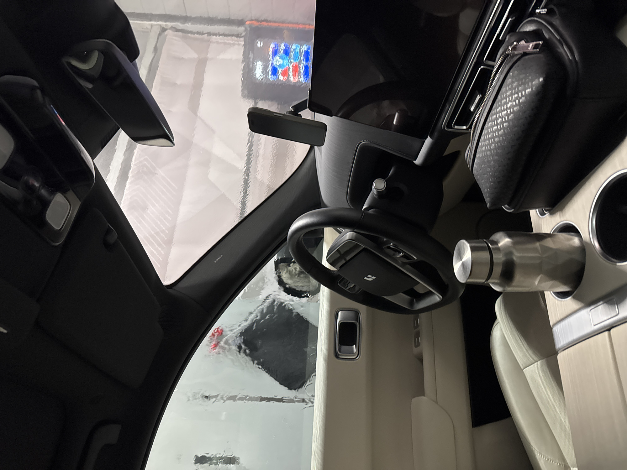 The image shows the interior of a car, possibly in a car wash, indicated by the water streaks on the windshield. The car has a black steering wheel with a square logo in the center. To the right of the steering wheel is a silver water bottle in a cup holder. A black zippered pouch is also visible. The interior is primarily black and beige. A visor is flipped down to the left, obscuring part of the view. A digital display is visible in the distance. The location is an unknown city.