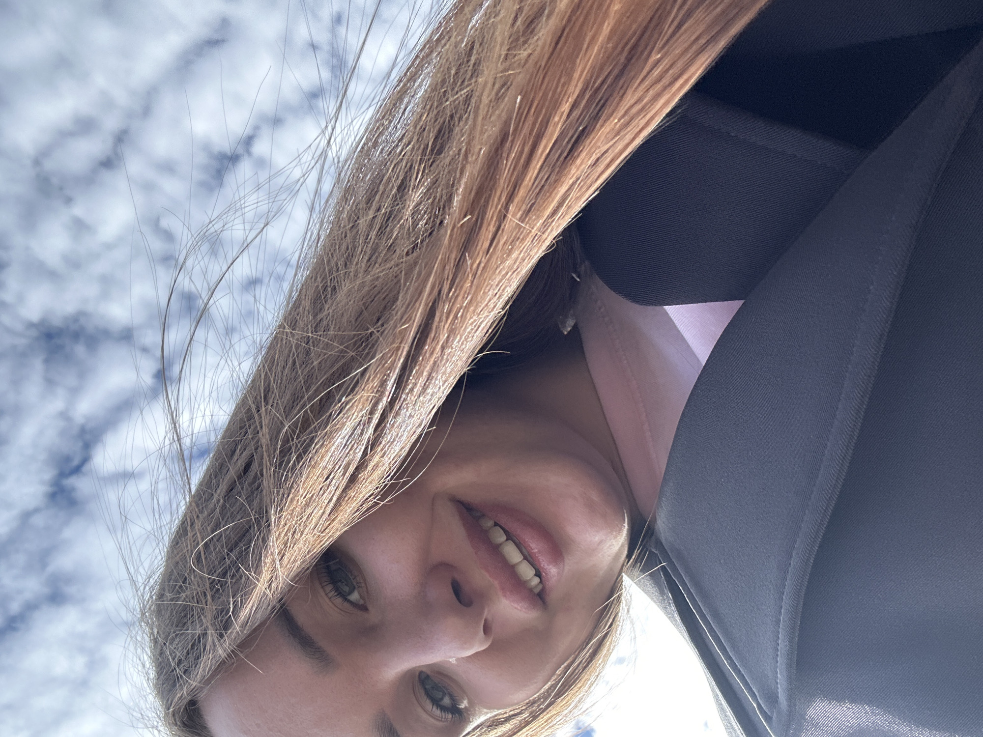 A low-angle, close-up outdoor portrait features a female individual in the city of Noviy, Russia. The subject is positioned against a background of a blue sky with scattered white cumulus clouds. Her long, light brown hair, appearing somewhat windblown, frames her face. She has green/hazel eyes and is looking directly towards the camera with a slight smile, revealing her upper teeth. She wears a light pink top beneath a dark gray or black outer garment, possibly a jacket or coat, with a visible structured lapel and collar. The immediate environment shows no ground-level features, only the expanse of the sky.