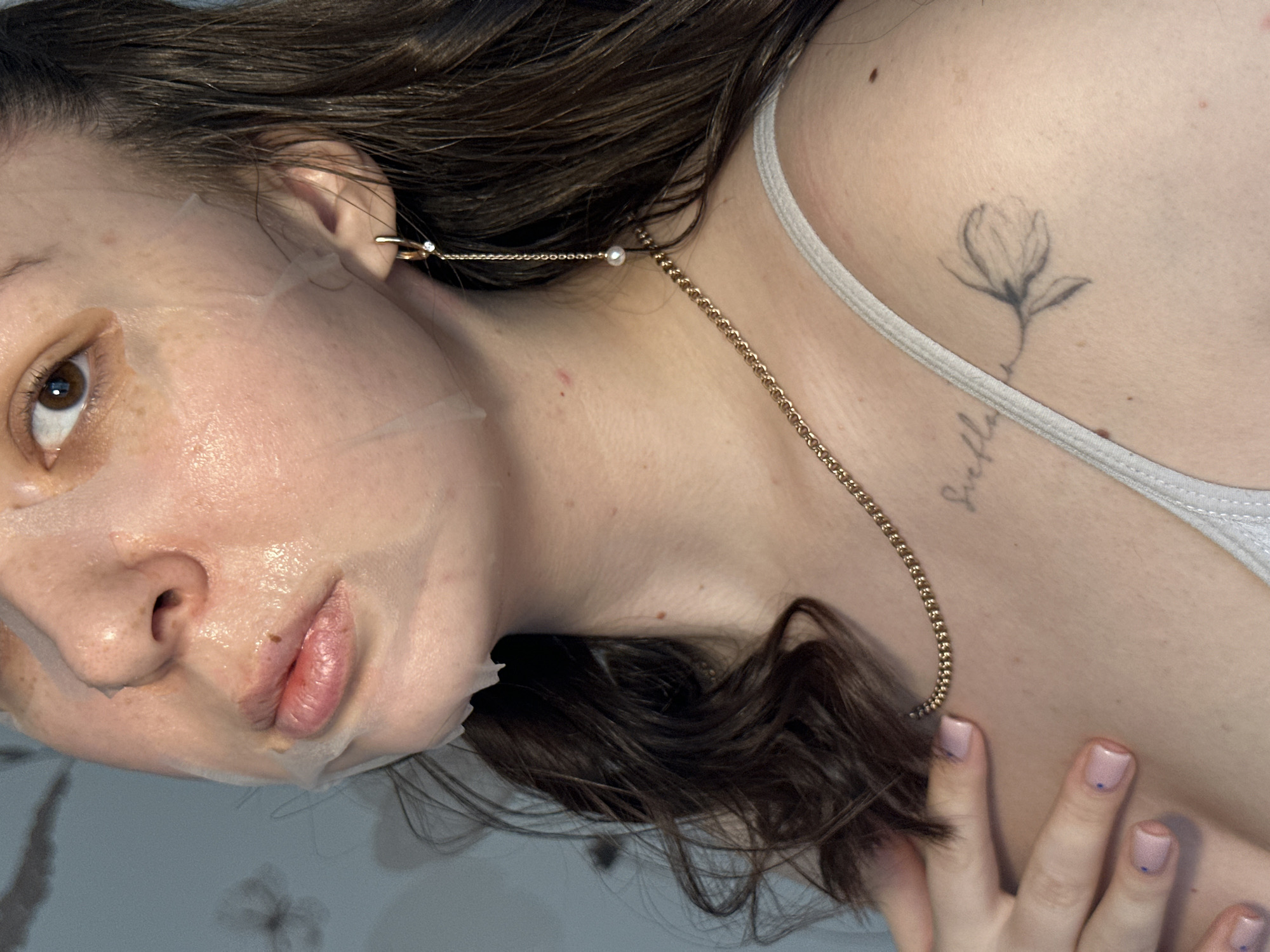 A woman is pictured indoors wearing a sheet mask on her face. She has brown hair and brown eyes, and her nails are manicured with small blue dots. She is wearing a gold chain necklace. Visible on her upper chest are tattoos of a flower and the word "Sickle." The image appears to be taken in a well-lit interior space with a light-colored surface beneath her. The location is not possible to determine from the image.