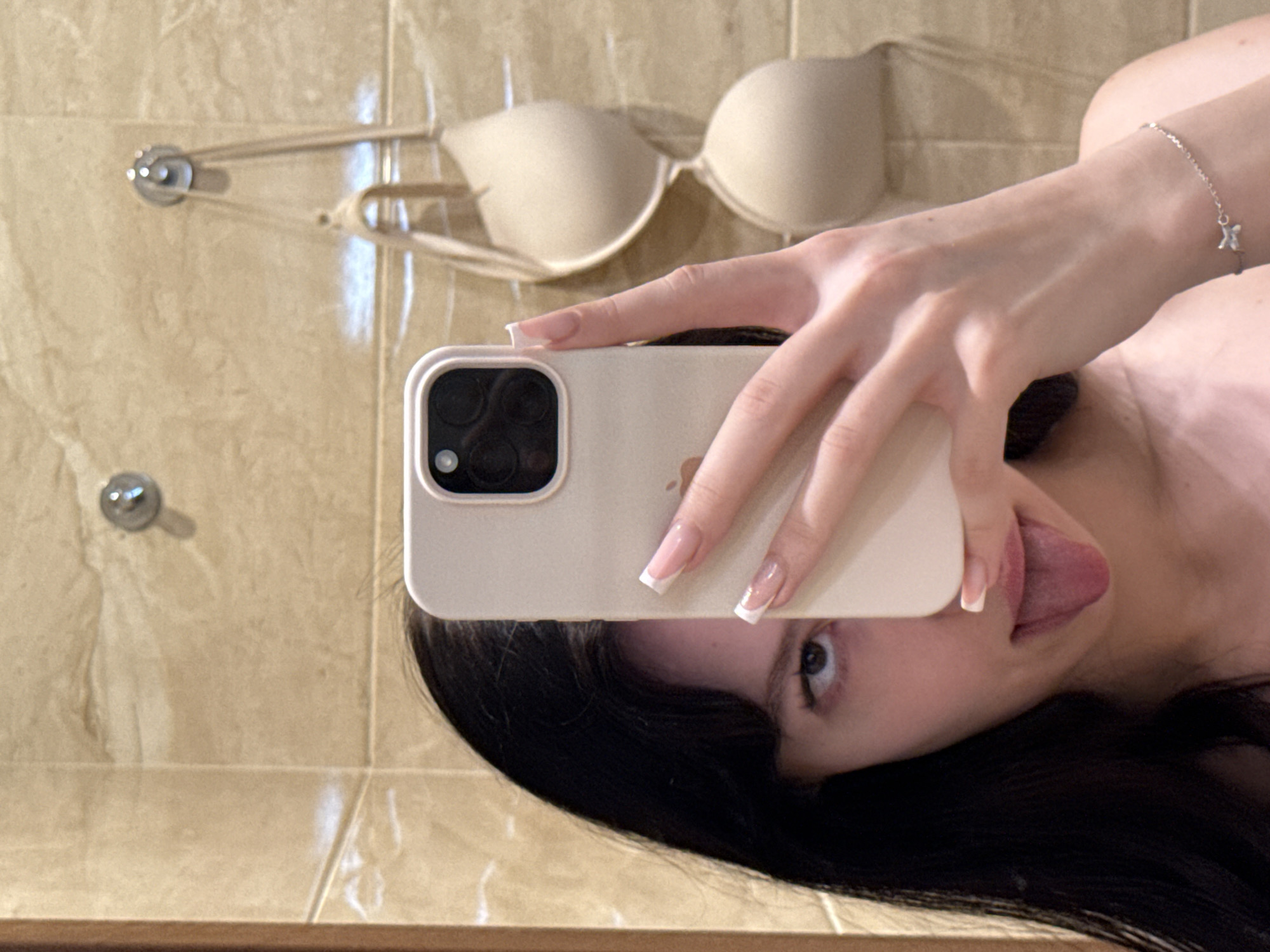 A young woman with long, dark hair and bangs is taking a selfie in a bathroom. She is holding a phone in her right hand, which is partially obscuring her face. She is sticking her tongue out, and her left eye is visible. She is wearing a silver bracelet on her left wrist. In the background, a beige bra is hanging on hooks on a tiled wall. The location is identified as the city of Khimki, Russia.