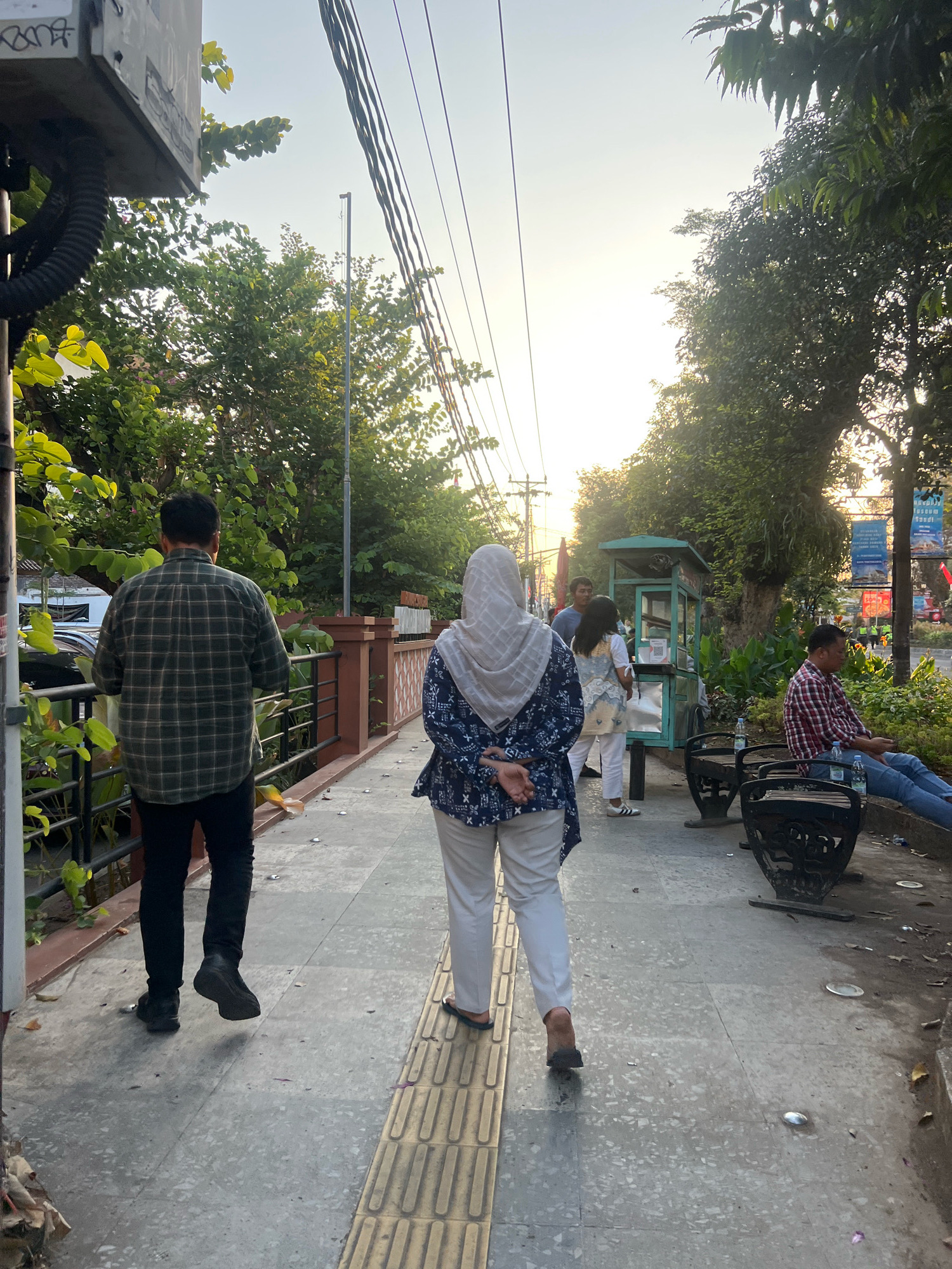 This image depicts a pedestrian walkway in Yogyakarta, Indonesia, likely near Malioboro Street, given the presence of street vendors and the general urban atmosphere. The time of day appears to be late afternoon or early evening, indicated by the warm, low-angled sunlight casting long shadows.
In the foreground, two individuals are walking away from the camera. The person on the left is a man wearing a dark green and black plaid long-sleeved shirt and black pants. The person on the right is a woman wearing a light-colored hijab and a blue and white patterned tunic over white pants.
Further down the walkway, several other people are visible. A vendor operates a small, green food cart. Two other individuals, a man and a woman, are standing near the cart, possibly interacting with the vendor. To the right, a man in a red and blue plaid shirt is sitting on a dark metal bench.
The environment includes lush green trees lining the walkway, overhead power lines, and decorative railings. The walkway itself is paved with light-colored tiles. Small, circular lights are embedded in the pavement. Debris, such as fallen leaves and wrappers, is scattered on the ground along the edge of the walkway. Banners with text are visible on poles in the background, suggesting a commercial or public area.