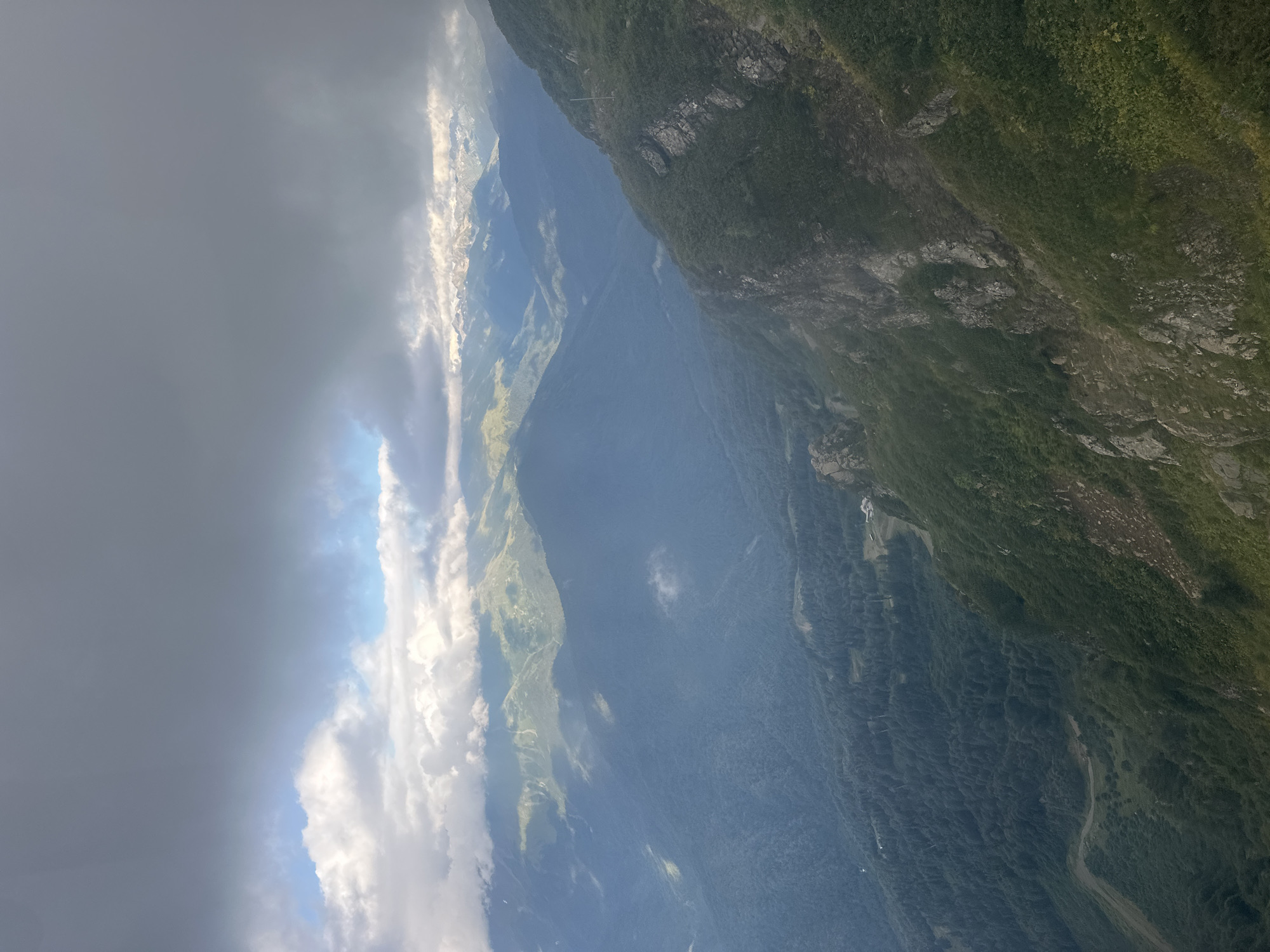 The image shows a view of mountainous terrain with a valley below, likely taken from a high vantage point. The sky is partly cloudy with dark storm clouds dominating the upper portion of the image and lighter, more scattered clouds below, allowing patches of sunlight to break through and illuminate the landscape. The mountains are covered in dense green vegetation, appearing as lush forests in the valley and on the lower slopes. The rugged, rocky slopes of the foreground mountain are covered in scrub and grass, with some exposed rock faces. In the distance, more mountain ranges are visible, some with snow-capped peaks, indicating a significant elevation. A few buildings, possibly houses or lodges, are visible nestled within the green landscape in the valley. The overall scene suggests a natural, remote, and possibly dramatic landscape, typical of mountainous regions during variable weather conditions. This view is characteristic of the natural landscape surrounding the city of Estosadok, Russia, which is known for its mountain resorts and natural beauty.