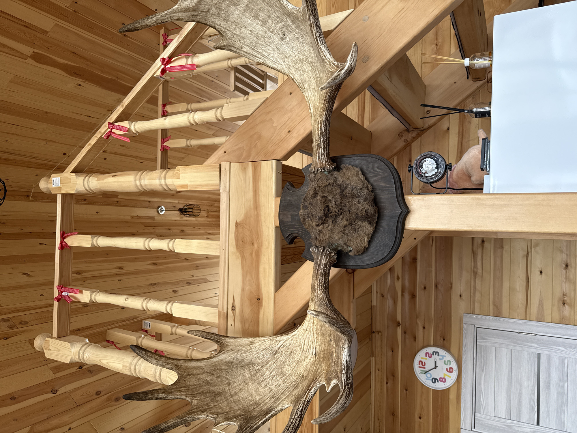 The image depicts an interior scene with prominent wooden elements. The space is characterized by walls clad in light-colored wood paneling, angled due to the building's structure. A staircase made of light-colored wood with balusters decorated with small red ribbons is present. A large moose antler rack is mounted on a dark, decorative wooden plaque, attached to a wooden beam structure. The scene also contains a wall-mounted clock with a white face and colorful numerals, and a gray door. Other objects such as an air freshener diffuser, disco ball, and part of a white refrigerator or cabinet can be seen in the background. There are no people explicitly visible in the image. The location is not evident from the image alone and would require additional context to determine if it is indeed Krasnoyarsk, Russia.