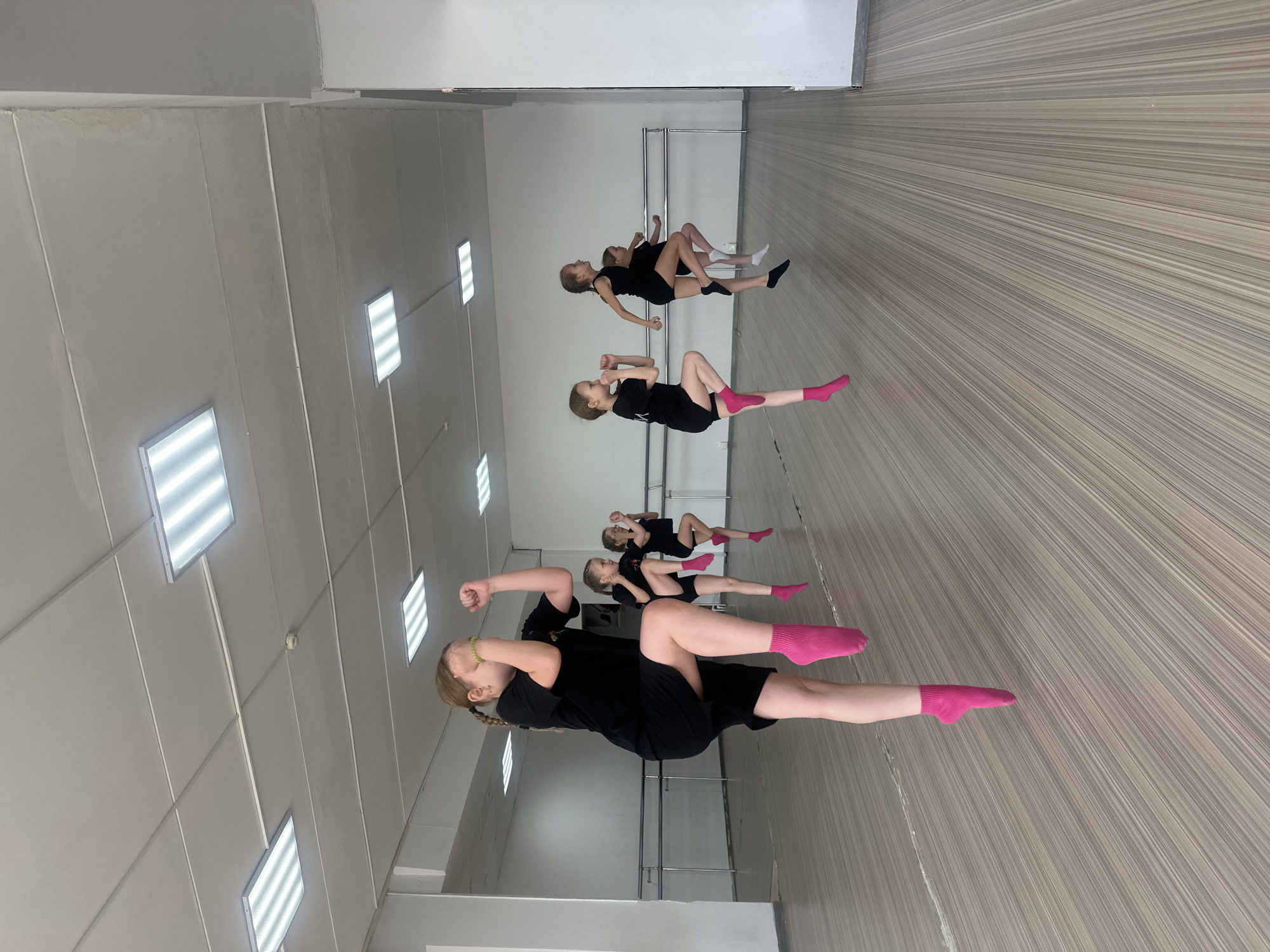 A group of girls are in a dance studio in Votkinsk, Russia. They are lined up performing a dance routine, with several girls visible. The dancers are wearing black dance attire. Some of them have pink socks. The dance studio has fluorescent lights on the ceiling, and a ballet barre along the wall. The floor has a linear pattern.