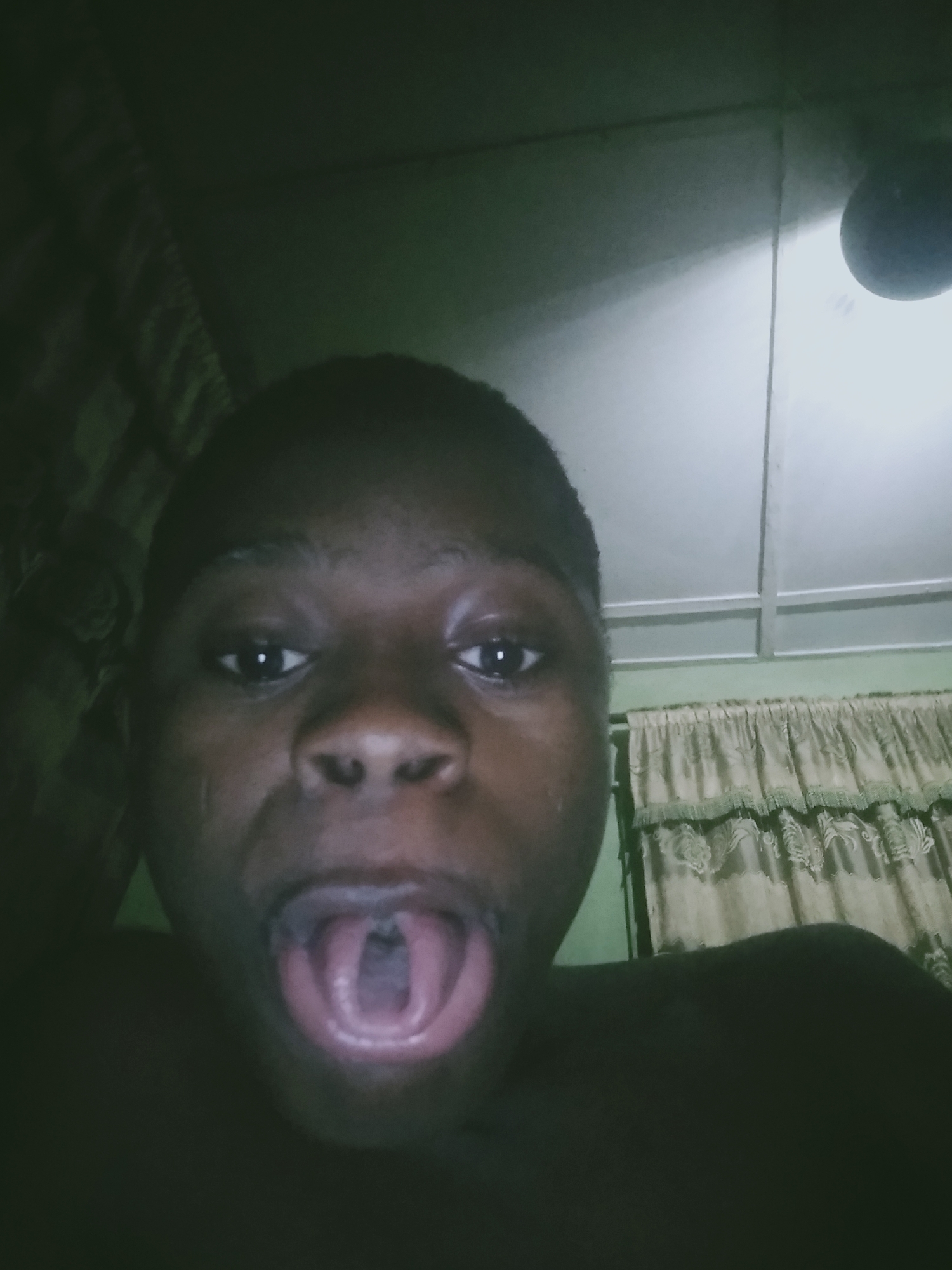 A person with dark skin is positioned close to the camera, facing forward with their mouth wide open, revealing the inside of their mouth. The person's expression appears exaggerated, with their eyes also wide.
Behind the person, the background reveals sections of a room. There is a light fixture visible near the top right corner of the frame, and curtain fabric is seen in the upper right quadrant. There is also a dark section to the left, possibly a curtain or a wall in shadow. The location of this image is in the city of Unknown, Unknown.