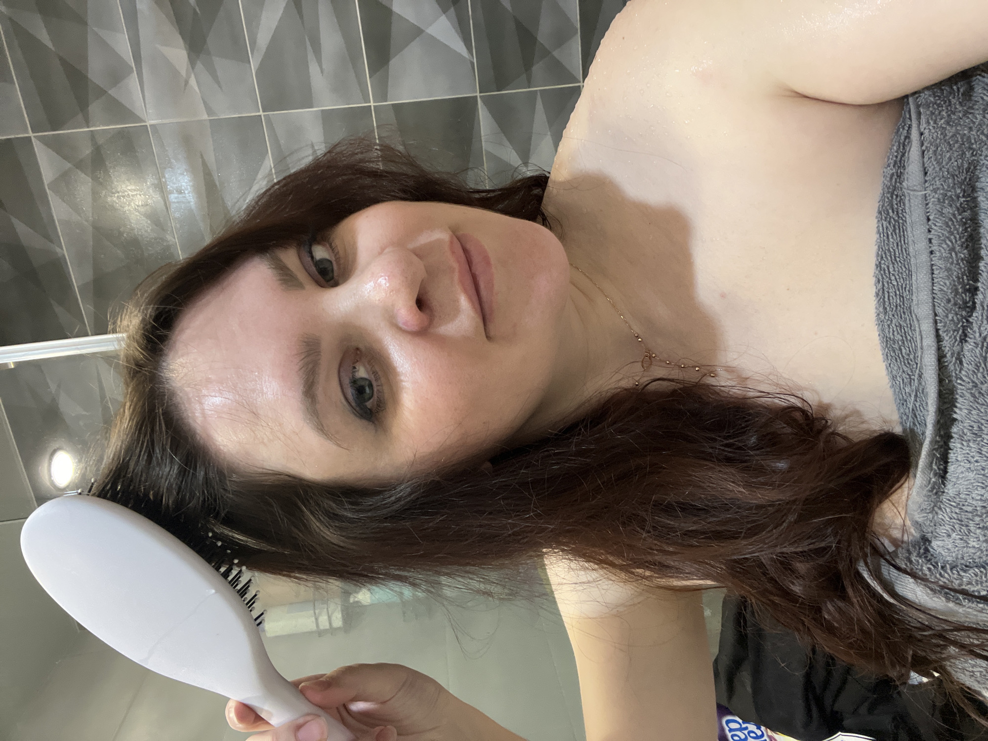 In Noviy, Russia, an adult female is depicted indoors within a bathroom environment. She possesses dark brown, wet hair and visibly moist skin with water droplets on her face and shoulders, suggesting a recent shower. She wears a grey terry cloth towel wrapped around her torso and a thin gold-colored necklace. Her right hand holds a white paddle brush, which is engaged with her wet hair on the upper left side of her head. She looks directly at the camera with a neutral expression. The background features a dark grey tiled wall with a repeating geometric pattern. A vertical metallic shower frame element is partially visible on the left, along with a reflection of an overhead light source. A dark object with partial white lettering is visible in the lower-left corner of the frame.