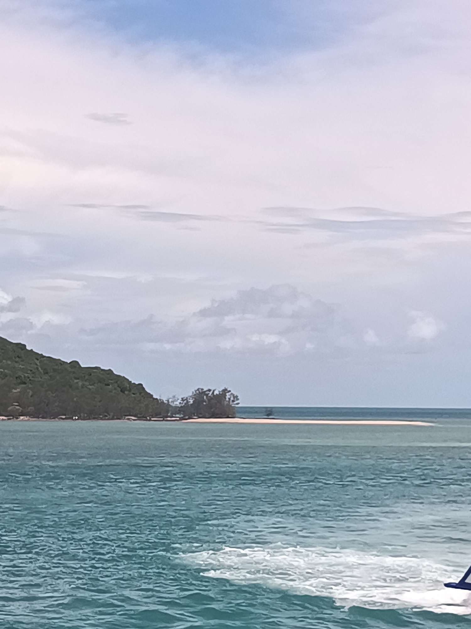 The image shows a coastal scene in Thong Sala, Thailand, likely taken from a boat moving on the water. In the foreground, there are ripples and white foam caused by the boat’s movement. The turquoise water stretches towards the horizon, where a long, narrow sandbar is visible. This sandbar is partially submerged and has a small patch of trees and vegetation in its center. To the left of the sandbar is a larger landmass, possibly an island, covered in dense, green vegetation. The sky is mostly cloudy with a mix of white and blue hues.
