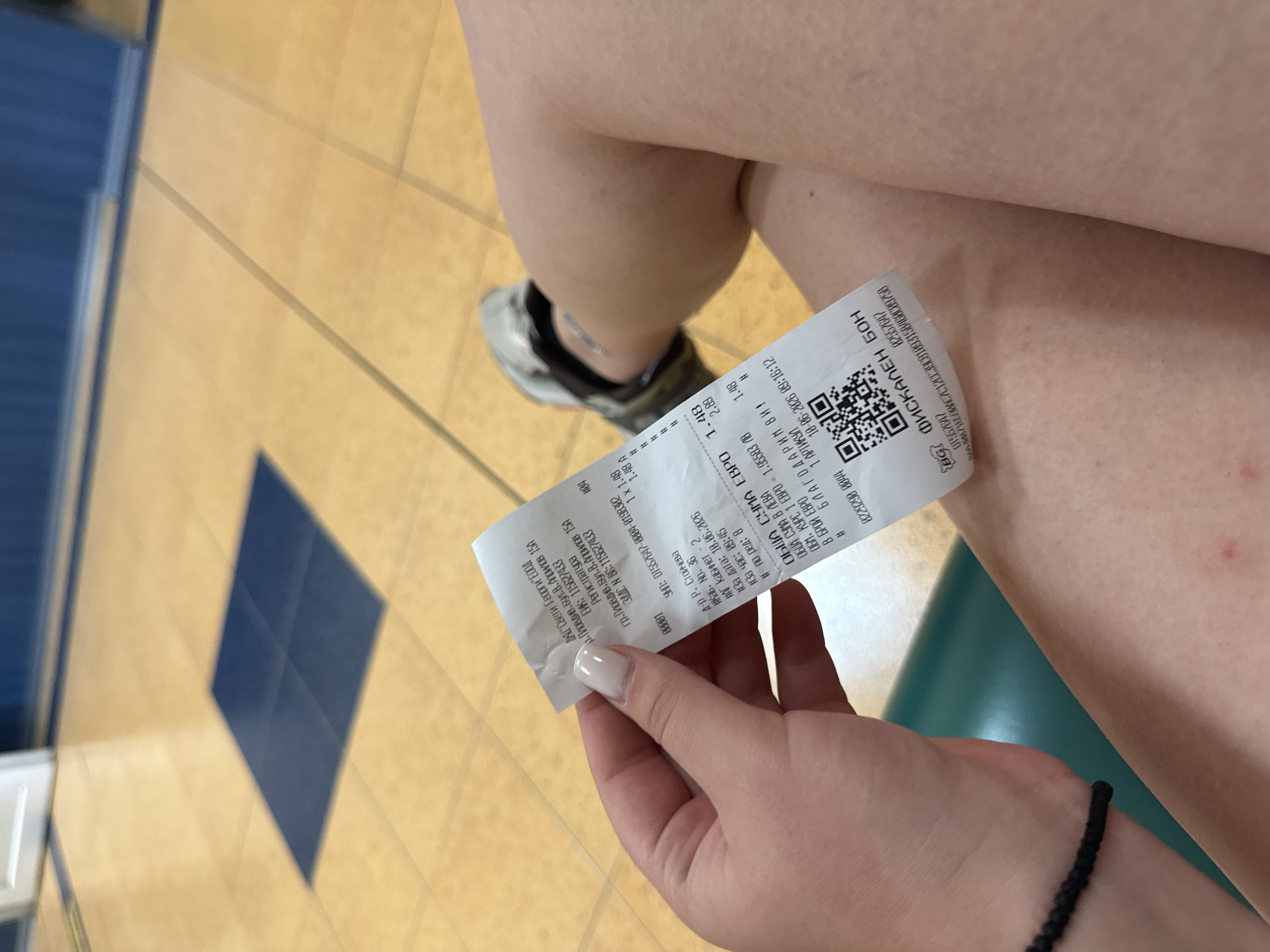 A person's hand, with a manicured nail, holds a crumpled receipt. The receipt is partially in focus and shows Cyrillic text and a QR code. The person's legs are visible, with one shoe on the floor. The background appears to be a tiled floor with blue square inlays and a blue wall. The receipt indicates the location as Plovdiv, Bulgaria, specifically mentioning "Пловдив, бул. В. Априлов 15A". This address corresponds to a commercial area, likely a shop or a service provider. The items listed on the receipt are in Euros, and the date is "18.06.2026". The scene is indoors, with artificial lighting.