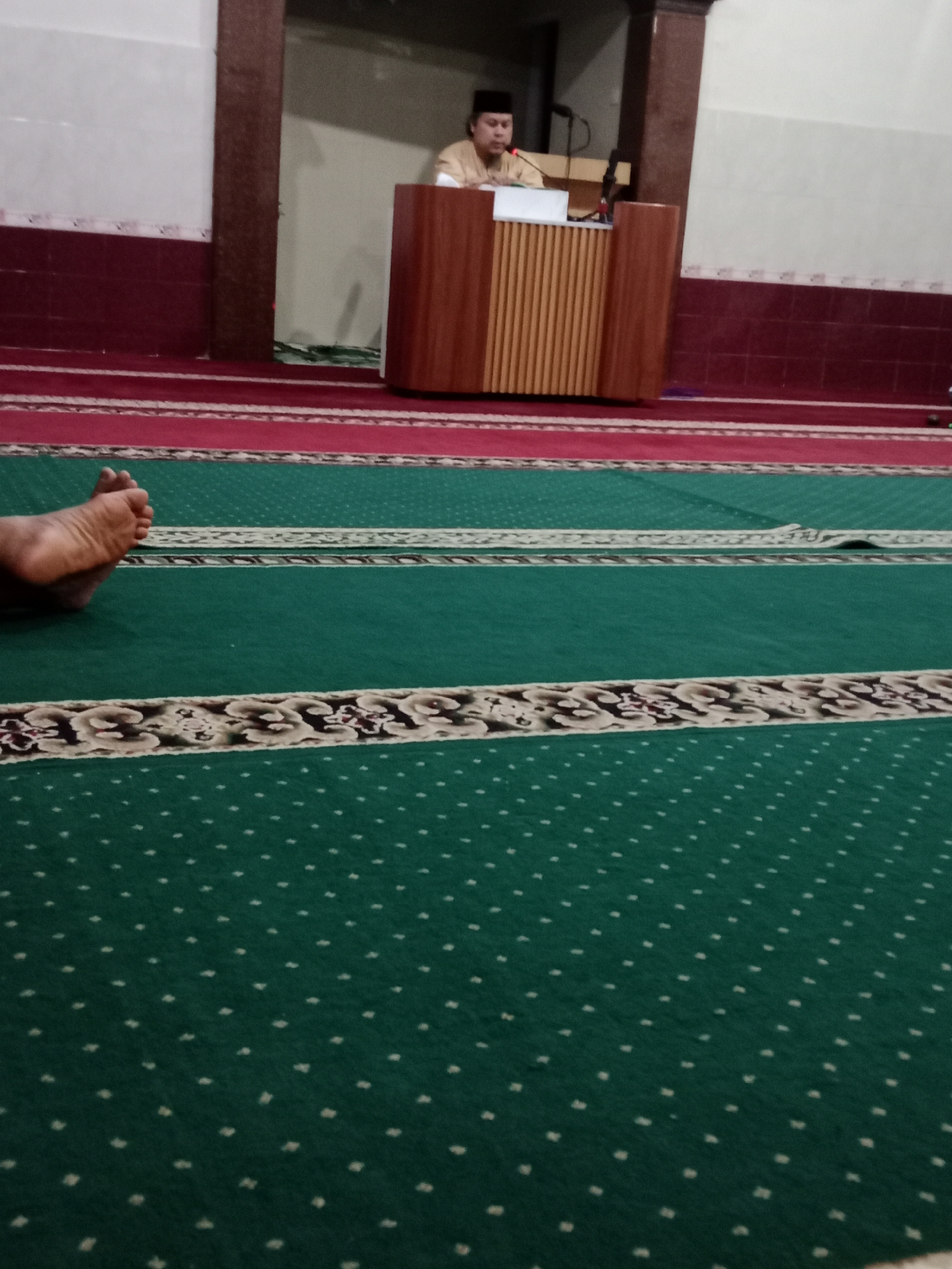 A man wearing a brown tunic and a white skullcap stands behind a wooden lectern with vertical slats. He is facing left and appears to be speaking or reading from a document. There is a microphone in front of him. The background shows a doorway with white walls and a dark wooden frame. The foreground is a green carpet with a dotted pattern and a decorative border. The red carpet is visible in the background, leading to the green carpet. A person's foot, with toes visible, is resting on the green carpet in the lower left corner. The scene appears to be inside a building, possibly a mosque or religious hall, given the carpet and the man's attire. There is no explicit indication of the city of Kediri, Indonesia, in the image itself.