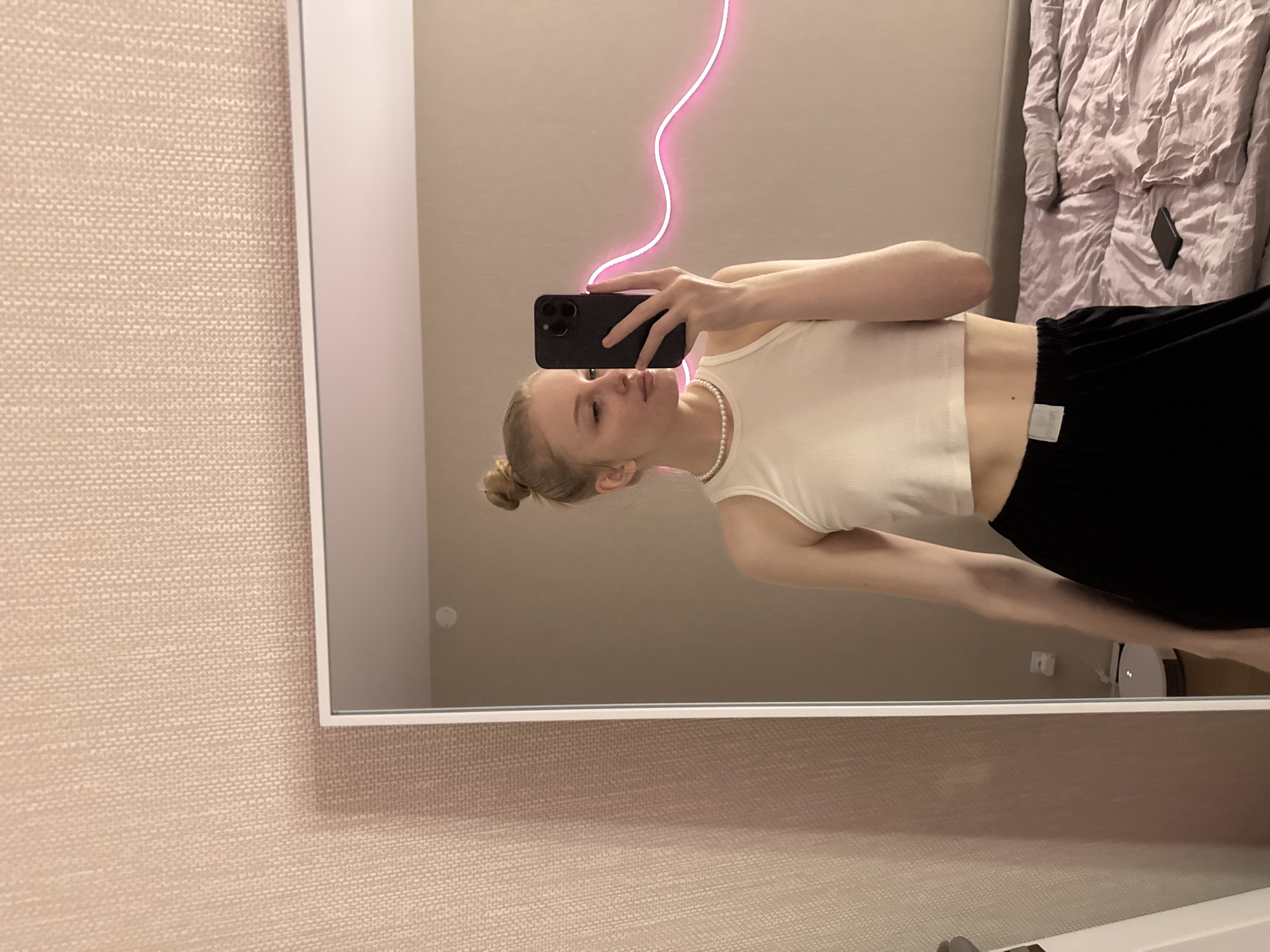 In Perm, Russia, a young blonde woman, with her hair in a messy bun, takes a mirror selfie. She wears a white ribbed crop top, a pearl necklace, and black loose-fitting pants, revealing her midriff. She holds a dark smartphone with a triple camera in her right hand, looking towards its reflection. The large mirror, framed in white, reflects a light beige wall behind her, adorned with a wavy pink neon light. A bed with crumpled light purple sheets and another dark smartphone lies in the reflected background. A white circular robot vacuum cleaner is visible on the floor in the reflection, beneath an electrical outlet. The wall outside the mirror's frame is covered in light beige textured wallpaper.