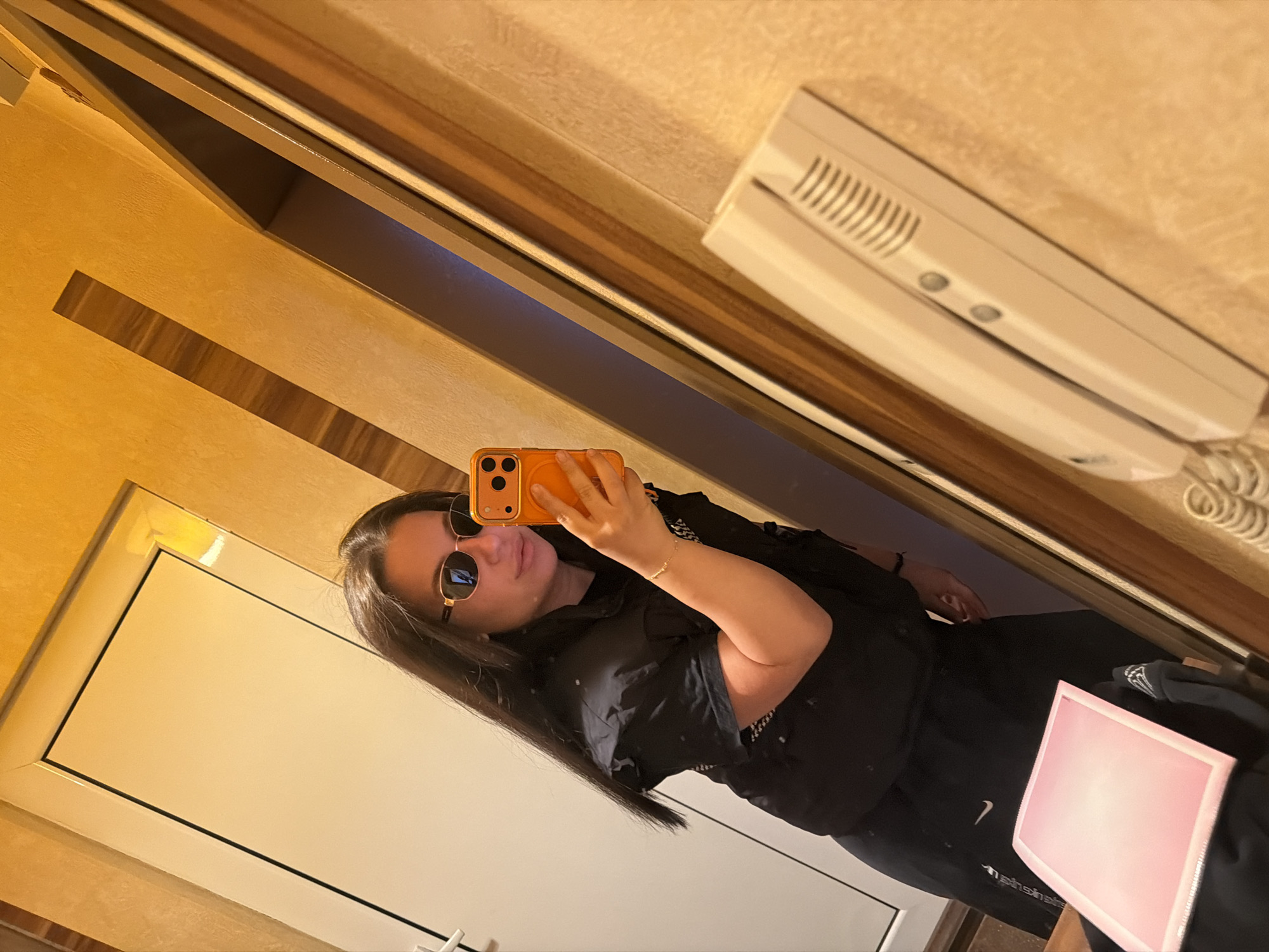 A person wearing sunglasses is taking a selfie in a mirror. The reflection shows part of an interior with a door, wooden-patterned walls, and an intercom system mounted on the wall. There is a pink rectangular object in the foreground near the bottom of the image. The location and city of Burgas, Bulgaria, cannot be determined from the image.