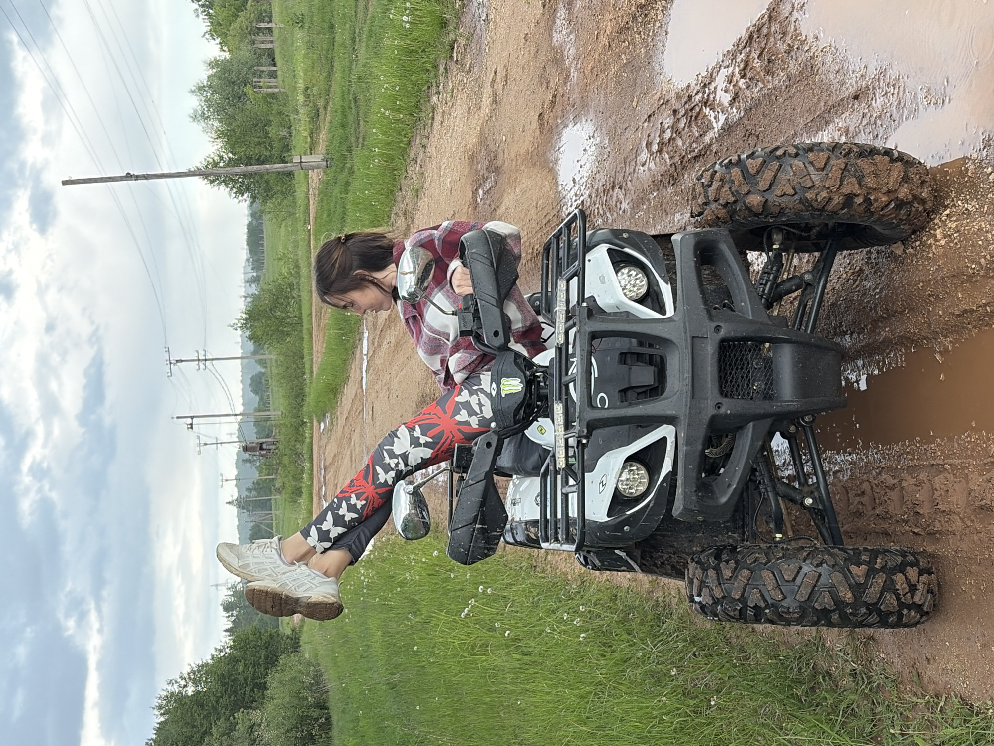 The image depicts a young woman seated on an all-terrain vehicle (ATV) in a muddy outdoor setting. The ATV is positioned on a dirt path with puddles, and the front tires are visibly coated in mud. The surrounding environment includes grass, trees, and a power line pole in the background. The woman is wearing a red and white plaid shirt, patterned leggings, and sneakers. She is holding the handlebars of the ATV, suggesting she has been riding or is about to ride. The scene is located in Nizhnii Vishur, Russia.