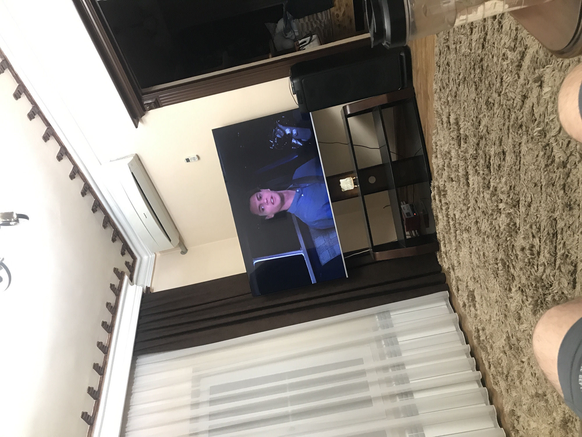 The image shows an interior scene, likely a living room. A television screen displays a man in a blue shirt. The TV is placed on a dark brown shelf unit. A large beige rug covers the floor. White curtains cover a window. An air conditioner is mounted high on the wall. Part of an upper window frame is visible. The location could potentially be in Yerevan, Armenia, but this is not definitively confirmed by the visual information.