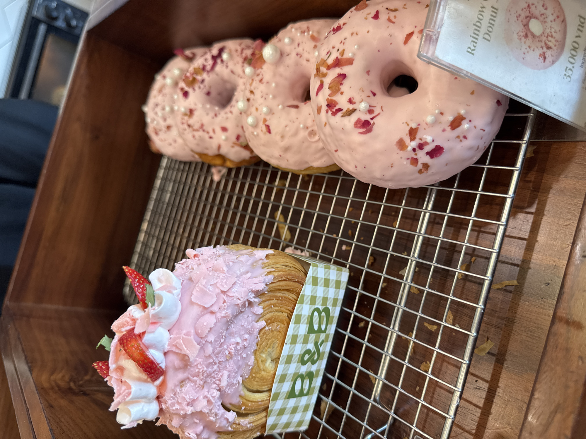 The image shows a display of pastries inside a wooden cabinet. On a metal wire rack, there are three pink-frosted doughnuts adorned with white pearl sprinkles and dried rose petals. A price tag indicates "Rainbow Donut - 35.000 vnd." Below the doughnuts, there is a pink-frosted croissant topped with whipped cream and sliced strawberries. The croissant is wrapped with a green-and-white checkered band labeled "B&B". The scene likely depicts a bakery or café in Đà Nẵng, Vietnam, given the price tag in Vietnamese Dong. There are no people visible in the frame.
