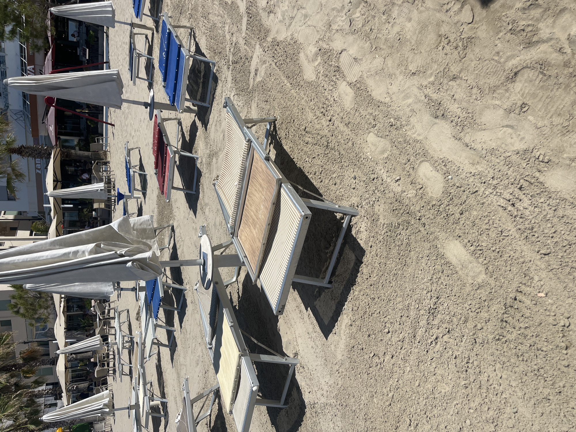 The image shows a beach in an unknown city. Rows of beach chairs and umbrellas are arranged on the sand. The sand appears to be tan colored. The chairs vary in color and style. Some have striped fabric seats and wooden frames, while others have blue or red fabric seats with metal frames. The umbrellas are mostly white. In the background, there is a building with a restaurant and palm trees. Footprints are visible in the sand.