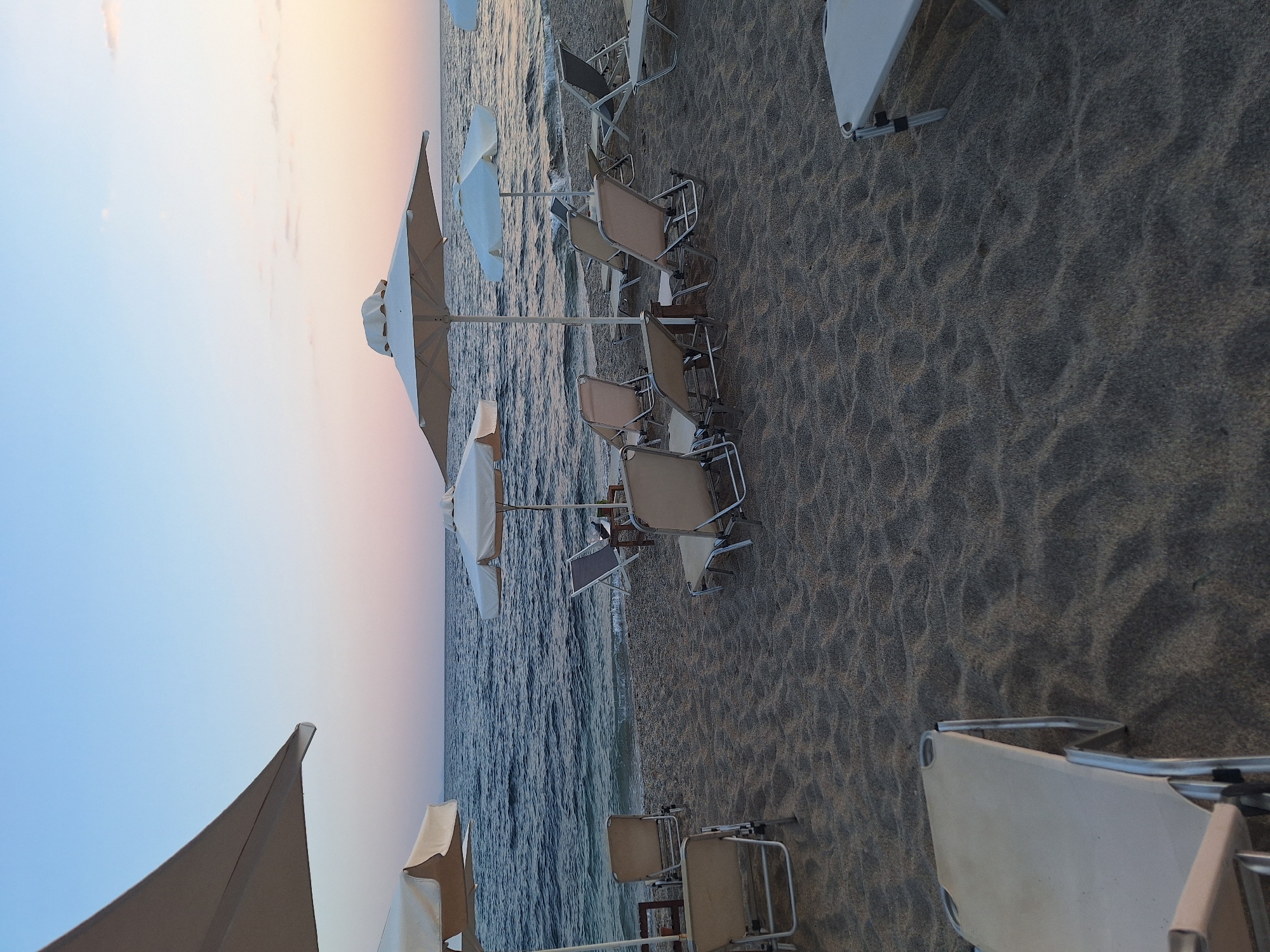 An empty beach at Mesimvria, Greece, is depicted in this image taken at dusk. The sky is a gradient of soft blues and pinks. The sea is dark with gentle waves reaching the shore. Several white beach umbrellas are open, casting shadows on the sand. Arranged beneath them are numerous lounge chairs, most of which are unoccupied. The sand is a fine, dark grain. In the foreground, partially visible lounge chairs and their shadows indicate the presence of more seating. The scene conveys a sense of calm and relaxation.