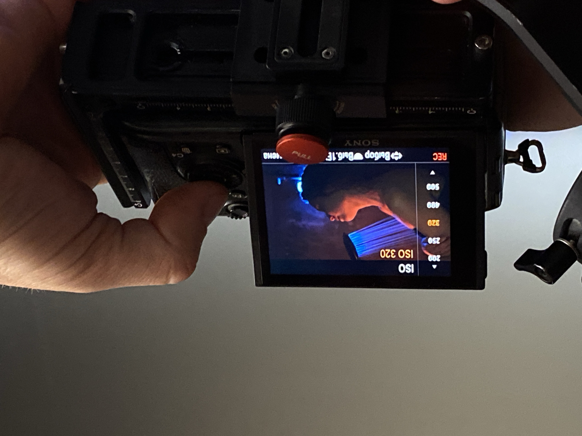 The image shows a close-up of a Sony camera, possibly a mirrorless or DSLR model, with the screen displaying a video recording. The ISO is set to 320. A person's hand is visible operating the camera's controls. A rig is attached to the top of the camera. The location is not indicated in the image.