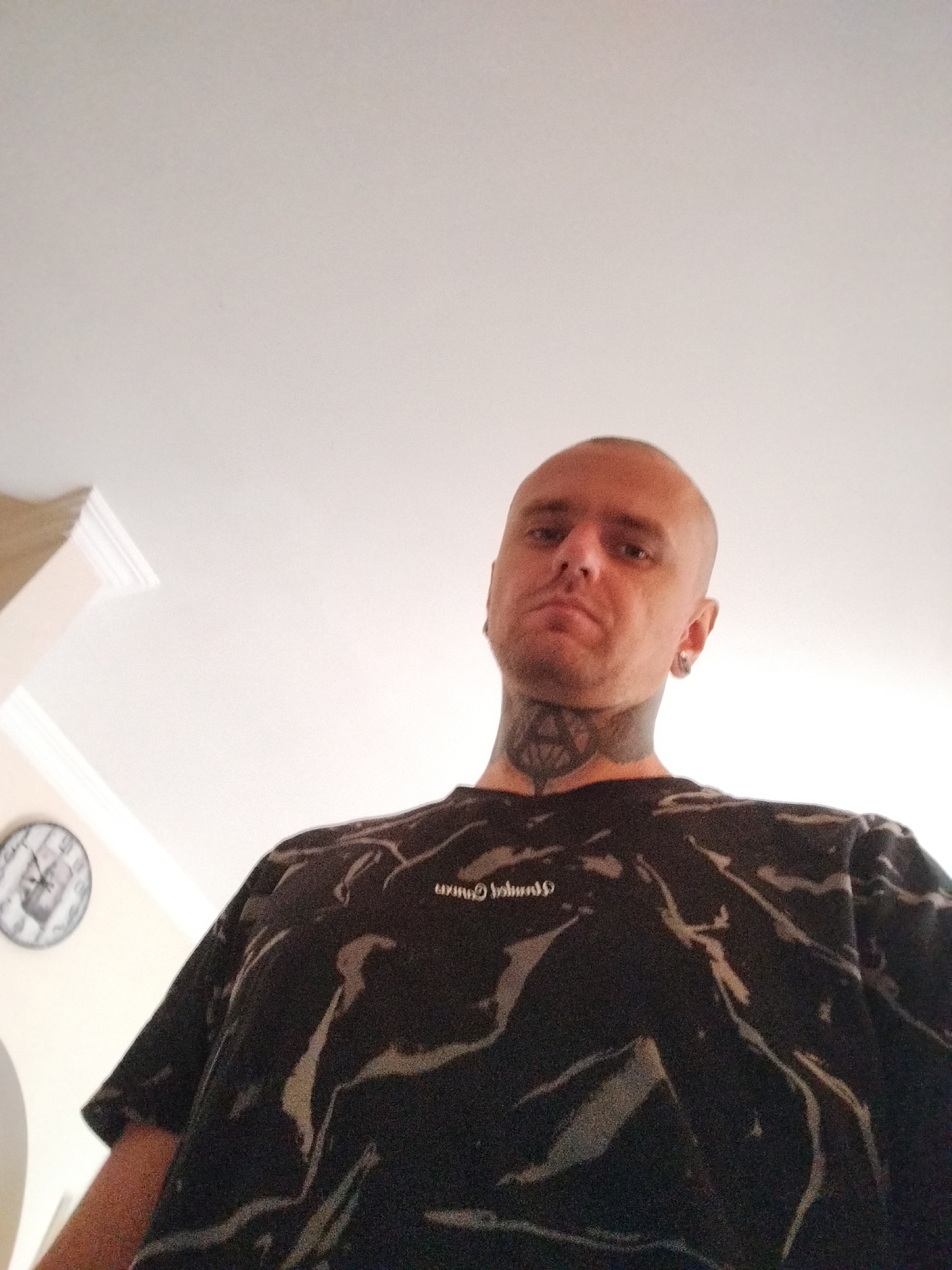 The image shows a low-angle shot of a bald man with tattoos on his neck. He is wearing a dark t-shirt with a marbled pattern. His head is tilted slightly down, and he is looking towards the camera. A wall with white molding and a clock are visible in the background. The lighting suggests an indoor environment, possibly a home. The presence of a clock on the wall could indicate a residential setting. There is no information to suggest this location is in Batumi, Georgia.