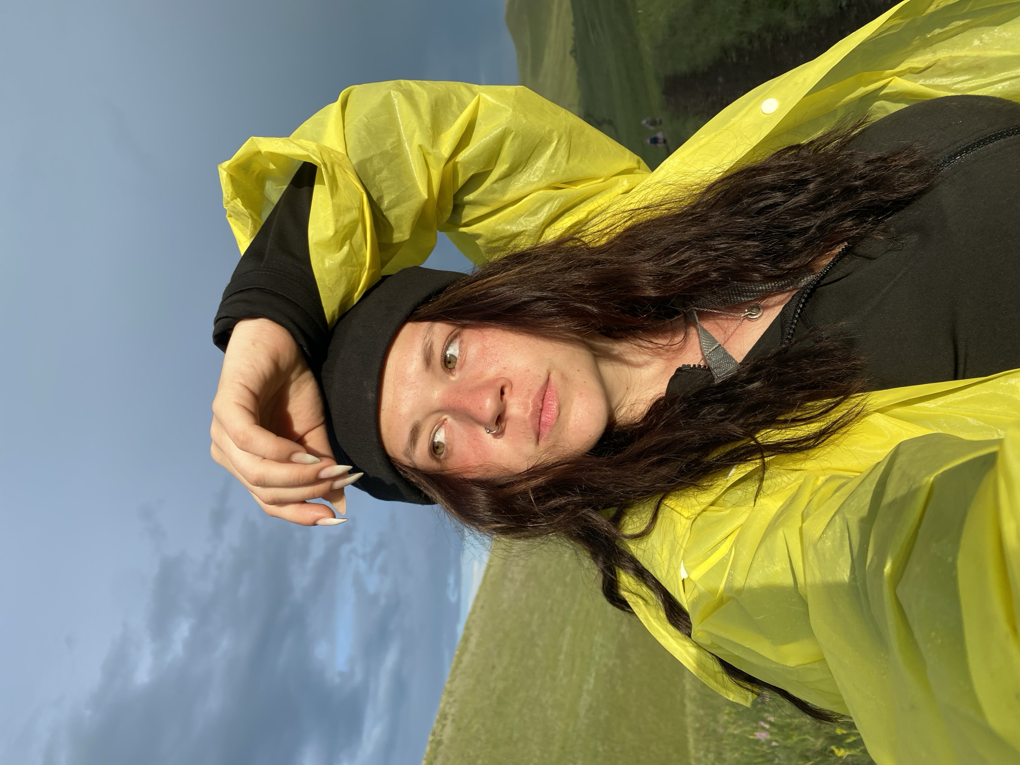 A woman, likely in her early 20s, is captured in a close-up, self-portrait style photograph against a backdrop of rolling green hills and a dramatic, cloudy sky. She is wearing a black beanie and a bright yellow raincoat. Her left arm is raised, with her hand resting on her forehead, and she is looking off to the right of the frame. Her long, dark, wavy hair frames her face, which features a nose ring and freckles. The lighting suggests it's either early morning or late afternoon, with a golden hue illuminating the scene. In the background, a dirt path winds up one of the hills, and two figures can be seen walking along it. The location is identified as Верхнеозёрное, Russia.